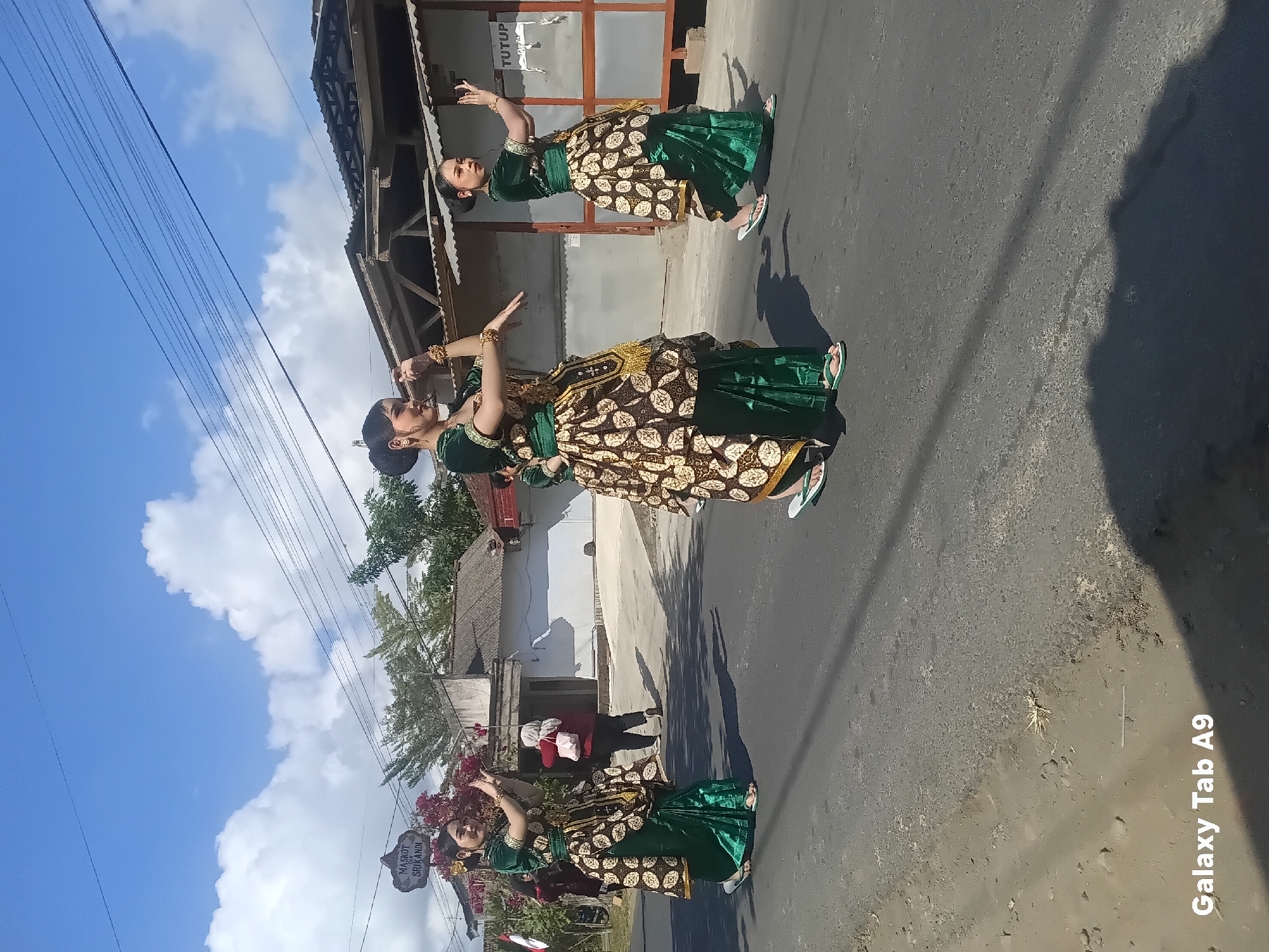 Three young women, adorned in traditional green and gold Indonesian attire, are parading down a paved road in what appears to be the city of Bendorejo, Indonesia. They are dressed in flowing green skirts, matching tops with intricate gold patterns, and decorated headpieces. Each woman wears gold bracelets on their wrists and bangles. Their hair is styled in elegant buns. The woman on the left is looking towards the camera, while the other two are looking forward. They are all in motion, with their arms positioned gracefully, suggesting they are dancing or performing.
Behind the women, a building with a corrugated metal roof and white walls is visible. A sign with red lettering that reads "TUTUP" (which means "closed" in Indonesian) is posted on the wall. Power lines stretch across the bright blue sky, which is dotted with fluffy white clouds. The road is asphalt and appears somewhat worn, with some debris scattered on the surface. In the background, there are trees and other buildings, indicating a residential or commercial area. The sunlight casts long shadows on the road, suggesting it is mid-day.