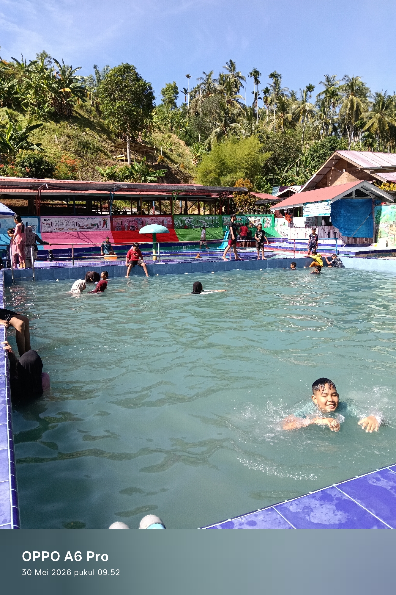 The image depicts a group of people swimming and playing in a pool at an outdoor recreational facility. The pool is bordered with blue tiles. Various people are visible inside the pool, including a boy swimming towards the camera in the foreground. Other individuals are standing at the pool's edge and swimming in the background. Behind the pool, there's a covered structure with colorful painted murals on the side. The setting appears to be in a rural or semi-rural area, with trees and vegetation covering a hillside in the background. Based on the timestamp, the photo was taken on May 30, 2026 at 09:52. The city is identified as Palohseulimeng, Indonesia.