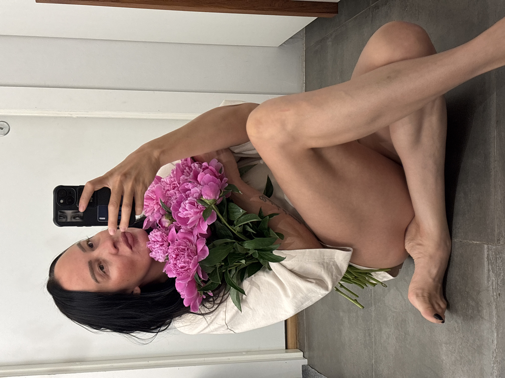 A woman with dark hair is lying on a gray tile floor next to a white wall. She is holding a black smartphone in her left hand, apparently taking a selfie. A large bouquet of pink peonies lies on her body. The background appears to be an indoor space, possibly a bathroom, with a wooden trim detail near the top of the wall. There is no indication that the location is in Chaikovsky, Russia.
