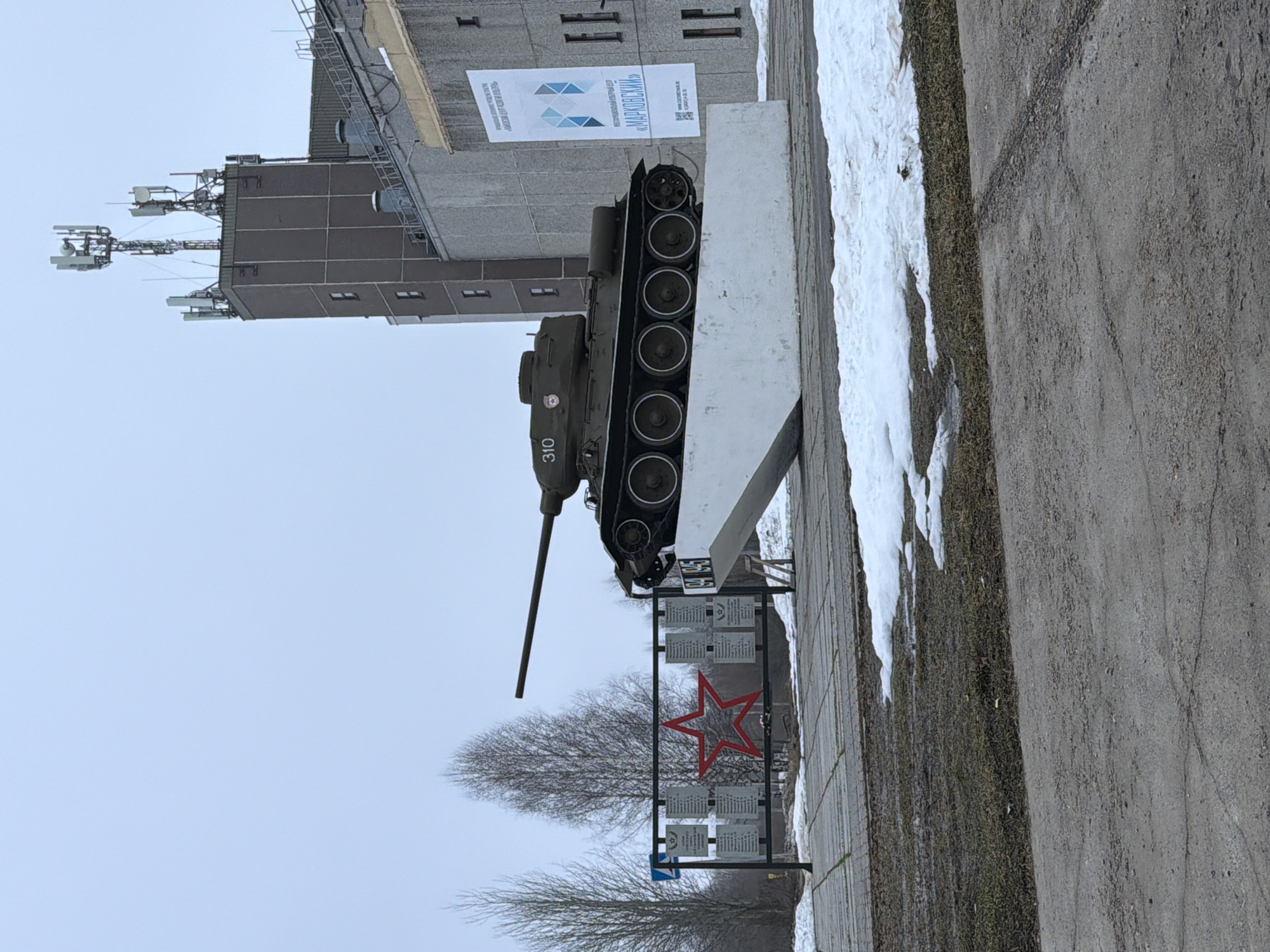 The image depicts a T-34 tank monument in Markovskiy, Russia. The tank is positioned atop a concrete structure. A sign that says "МАРКОВСКИЙ" is present on a nearby building. A red star is displayed on a metal frame in front of the tank. Snow is visible on the ground next to the tank, and a paved area and some grass are in the foreground. The sky is overcast. Buildings with antennas are visible in the background.
