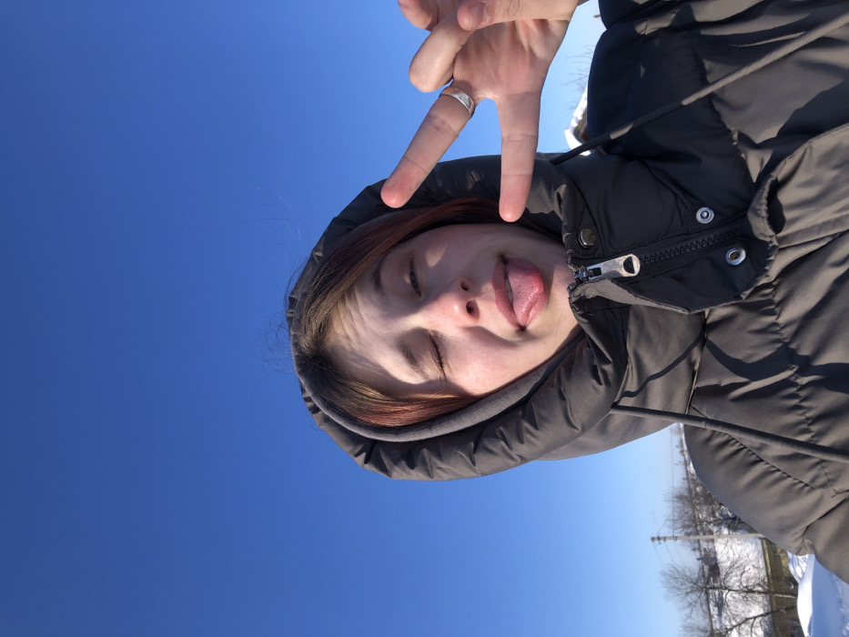 An image depicts a close-up of an individual, likely female, against a clear blue sky and snow-covered landscape in Фоки, Russia. The subject is wearing a dark grey hooded puffer jacket with the hood up, partially obscuring reddish-brown hair. Her right eye is completely closed, her left eye is squinted, and her tongue is extended from her mouth. Her left hand (viewer's right) is raised, with the index and middle fingers extended in a V-shape, and a silver-colored ring is present on the ring finger. The background features a bright, cloudless blue sky occupying the upper third of the frame. Below the sky, snow-covered ground is visible, along with bare deciduous trees and a utility pole on the left. Faint outlines of distant snow-covered structures are discernible on the horizon.