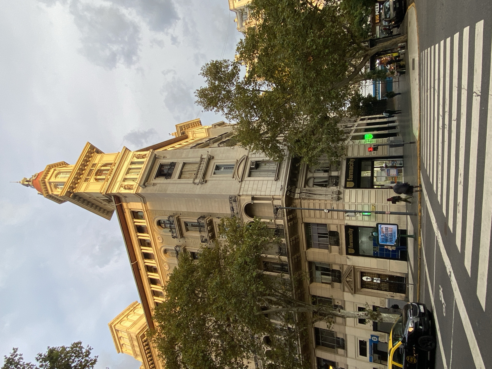 A street-level view of Avenida Roque Sáenz Peña (Diagonal Norte) in Buenos Aires, Argentina, displays a tall, ornate building with a distinctive red-domed tower on its upper left. The building is beige and features numerous windows, balconies, and architectural details like columns and molding. Green trees partially obscure the building's facade.
On the right, a zebra crossing marks the street's edge, leading towards storefronts. A "farmacity" pharmacy is visible. A digital advertising board displays "Con la Universidad Pública". A black and yellow taxi is parked at the curb. Pedestrians are walking on the sidewalk near the pharmacy. The sky is overcast with grey clouds.