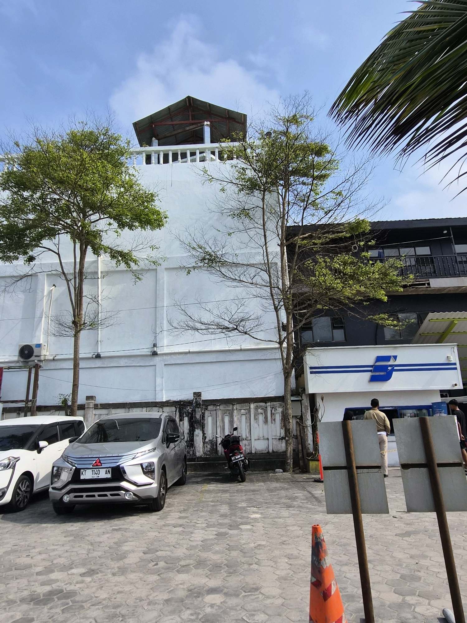 The image depicts an outdoor scene, likely a parking area, in Balikpapan, Indonesia. In the foreground, a silver car is parked on a paved ground made of hexagonal stones. To its left, a white car is partially visible. Behind the cars, a red motorcycle is parked.
The background features a white building with a balcony at the top, which has a dark, angular roof structure. Adjacent to this building is a darker, multi-story structure with windows and balconies. A white building with blue stripes and a blue logo, possibly a business or transport company, is also visible.
Several trees are present, some with green foliage and others appearing bare. A palm frond extends from the top right corner of the image. Two men are standing near the building with the blue logo, one facing away from the camera. Two weathered metal signs on wooden posts stand in the mid-ground, and an orange traffic cone is in the lower right. The sky above is a bright blue with scattered clouds. The presence of the license plate "KT 1140 AN" on the silver car, where "KT" is the vehicle registration code for East Kalimantan, confirms the location as Balikpapan, Indonesia.