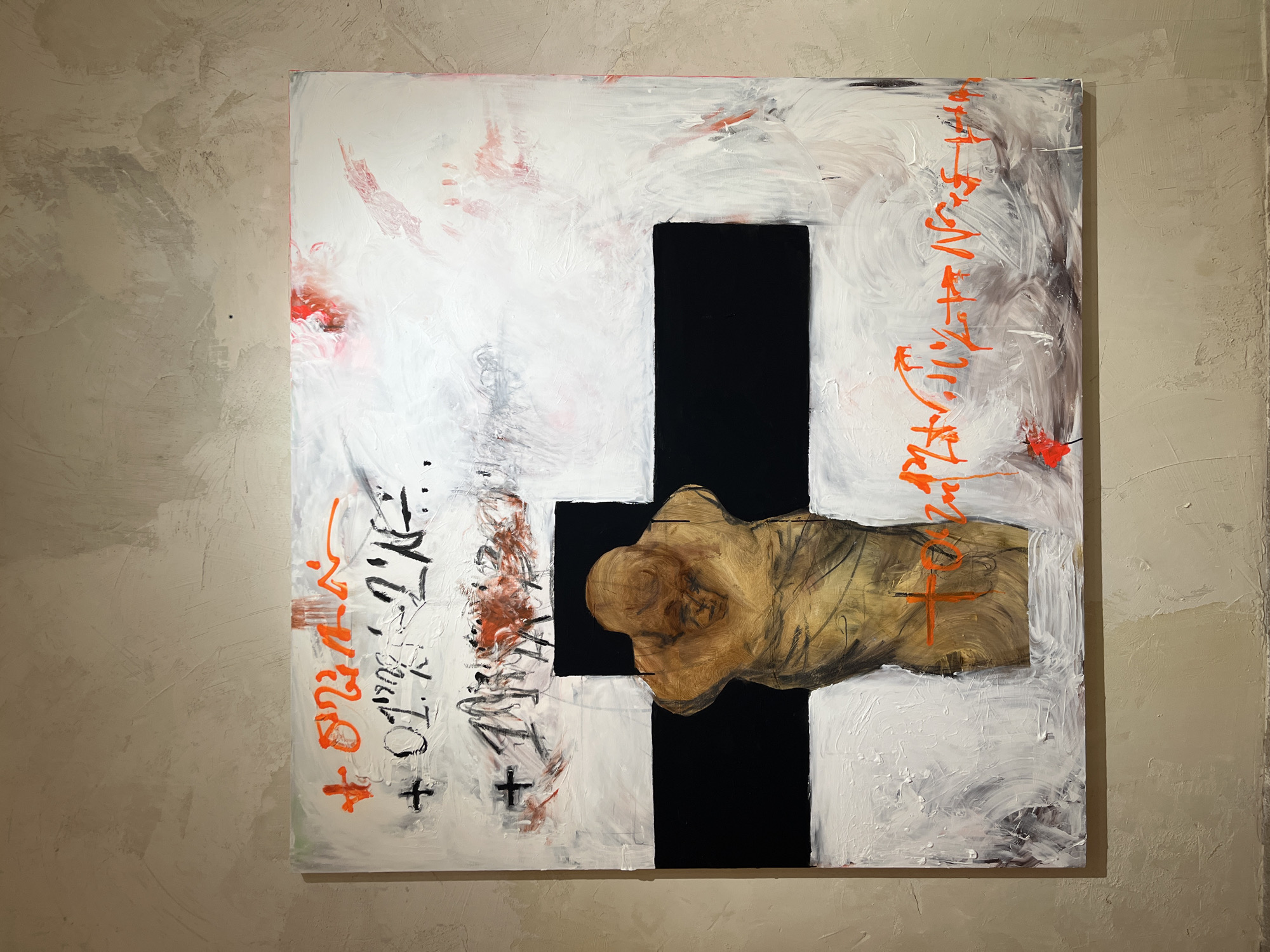This is an abstract painting featuring a large, dark cross against a white background with various textures and markings. A piece of wood, resembling a human torso or torso-like form, is affixed to the center of the cross. The artwork is displayed on a textured, beige wall, suggesting an indoor setting like a gallery or studio.
The white background is textured with thick brushstrokes, some appearing rough and others smoother. There are areas of red and black paint splattered and smeared across the white surface, adding to the abstract and somewhat chaotic feel. Orange, calligraphic-like markings are visible on both the left and right sides of the painting, appearing to be writing or abstract symbols. Some of these orange markings form what look like cross shapes as well. A few black cross shapes are also present on the lower left portion of the canvas. The wood element is a prominent feature, with visible grain and texture, and it is rendered in natural wood tones with some darker lines suggesting form or shadow. The overall impression is one of intensity and emotional depth, perhaps exploring themes of spirituality, sacrifice, or the human form. The lighting is dim, casting soft shadows and highlighting the textures of both the painting and the wall. The style is mixed media, combining painting with sculptural elements.
