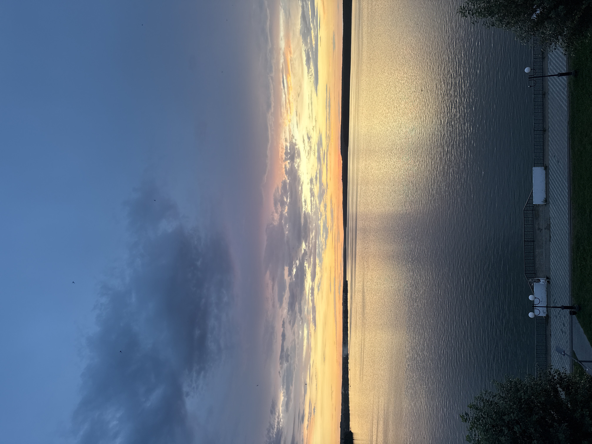 The image captures a scenic view of the Votkinsk Reservoir in Votkinsk, Russia, during twilight. The sky is a canvas of deep blues and purples, transitioning into vibrant oranges and yellows near the horizon, indicative of a sunset or sunrise. Wispy clouds are scattered across the sky, with a few birds visible as small dark specks. The reservoir itself is calm, reflecting the colors of the sky and creating a shimmering effect on its surface.
In the foreground, a paved walkway runs along the edge of the reservoir, bordered by a low fence. A lamppost with two spherical lights stands on the walkway, and white benches are placed along the promenade. Green grass and trees can be seen beyond the walkway, suggesting a park or public area. The distant shoreline is lined with trees, forming a dark silhouette against the colorful sky. The overall impression is one of tranquility and natural beauty.
