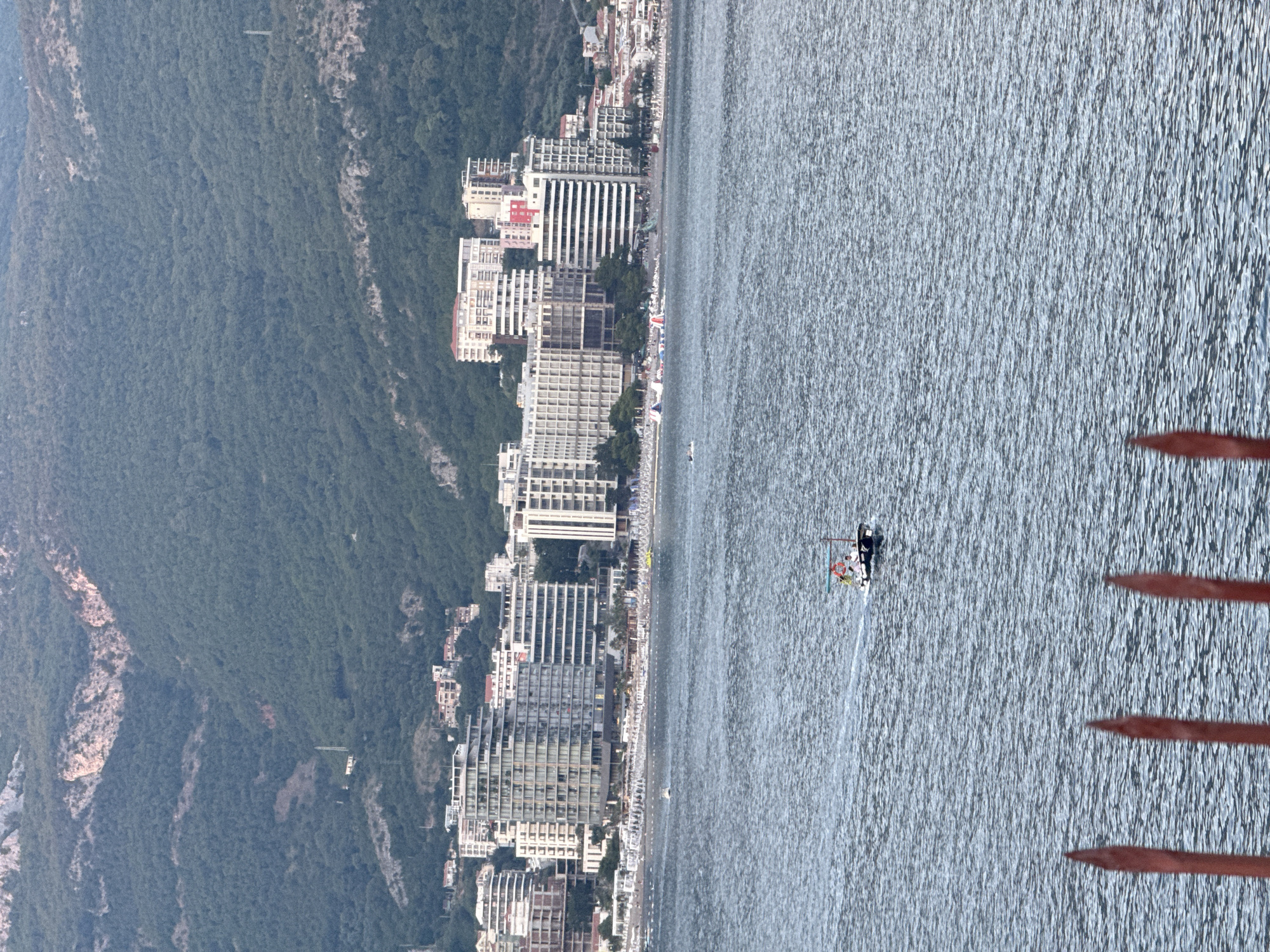 The image depicts a coastal scene in Budva, Montenegro. In the foreground, the calm, blue water of the Adriatic Sea fills the lower portion of the frame, with ripples reflecting the light. A small boat with a few passengers is visible in the middle distance, moving across the water. In the background, a densely populated coastline is lined with numerous multi-story buildings, predominantly hotels and apartment complexes, with their modern architecture contrasting against the natural landscape. Beyond the buildings, a steep, verdant mountain rises dramatically, covered in dense green foliage. The sky is overcast, contributing to the soft, diffused light that bathes the scene. The overall impression is one of a bustling coastal resort town nestled between the sea and the mountains.
