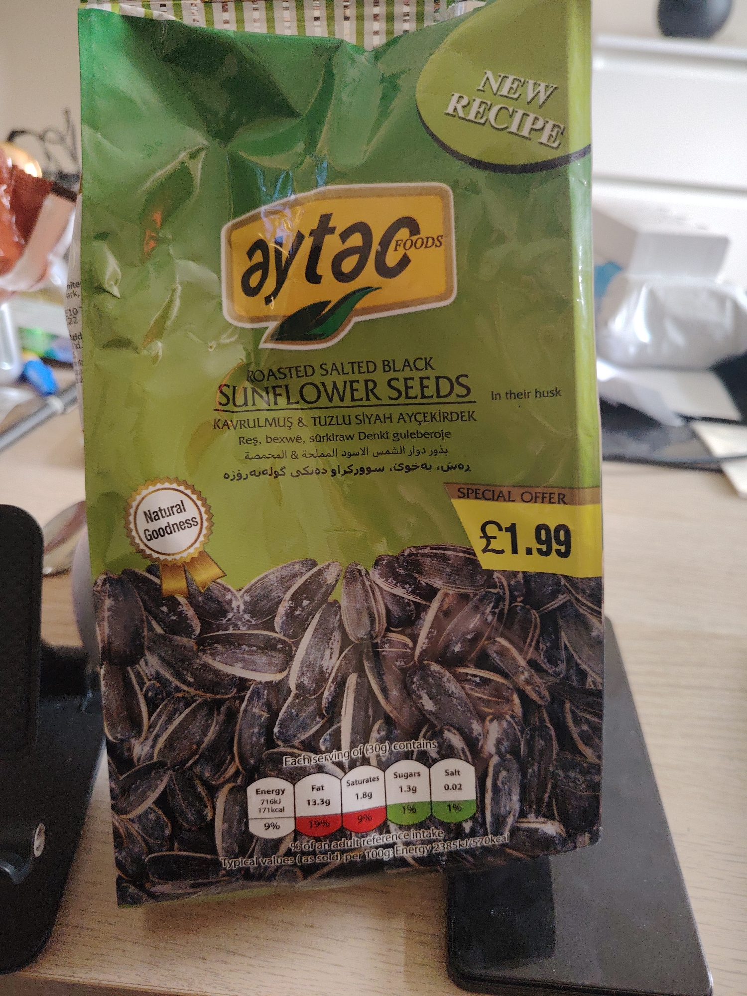 A green, branded plastic bag of Aytac Foods Roasted Salted Black Sunflower Seeds is prominently displayed in an indoor, well-lit setting. The packaging features the brand name "aytæc FOODS" in a yellow and black logo near the top. Beneath this, the product is identified as "ROASTED SALTED BLACK SUNFLOWER SEEDS" with the phrase "In their husk" to the right. Multilingual text is visible, including "KAVRULMUŞ & TUZLU SİYAH AYÇEKİRDEK" (Turkish), "Reş, bexwê, sûrkiraw Denkî guleberoje", "بذور دوار الشمس الاسود المملحة & المحمصة", and "ره ش به خوی سوورکراو ده نکی گوله به روژه" (Arabic and Kurdish).
Notable marketing details include a "NEW RECIPE" badge in the upper right corner and a "Natural Goodness" ribbon graphic on the left side of the bag. A "SPECIAL OFFER £1.99" price tag is visible on the right, indicating a price in British Pounds. The bottom section of the bag showcases a large image of the black sunflower seeds in their shells.
Below the seed image, a nutritional information table details the values for "Each serving of (30g) contains": Energy (716kJ/171kcal, 9% of reference intake), Fat (13.3g, 19%), Saturates (1.8g, 9%), Sugars (1.3g, 1%), and Salt (0.02g, 1%). The "Typical values (as sold) per 100g" are listed as "Energy 2385kJ/570kcal". The bag appears to be resting on a dark, flat surface, possibly a weighing scale or a counter. The background is blurred, showing hints of other household items, suggesting a domestic environment in Huntingdonshire, United Kingdom.