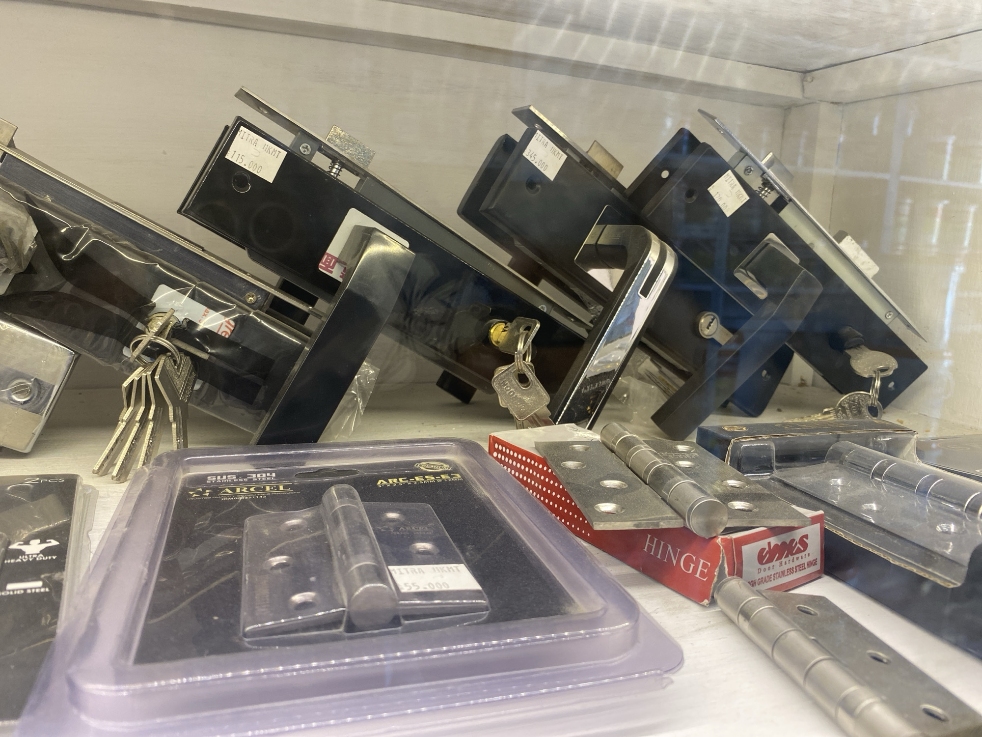 The image shows a display shelf in a store, likely a hardware store, with various door hardware items. In the foreground, there are packaged door hinges and what appear to be door locksets with keys. The locksets are mostly black and silver, with some featuring labels with prices. One label clearly reads "¥115.000" and another "¥345.000". Towards the back of the shelf, more locksets are visible, some with keys attached. The environment appears to be indoors with artificial lighting, creating reflections on the glass or plastic packaging of the products. The overall impression is that of a retail display of door hardware.