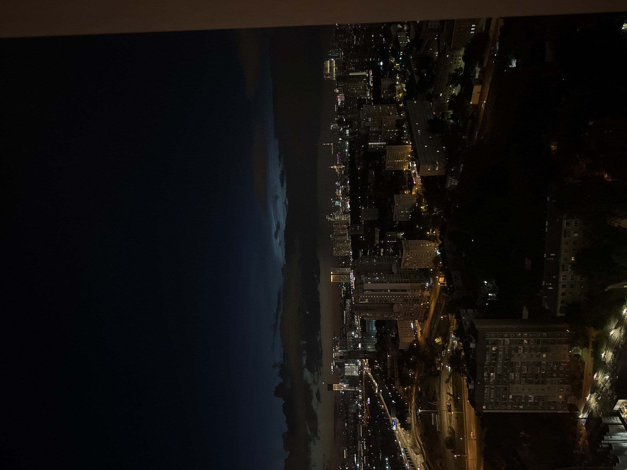 The image shows a panoramic nighttime aerial view of a city. The scene is divided into two distinct halves: the left side is dominated by a dark, cloudy sky, suggesting a storm or impending rain. The right side of the image reveals a sprawling cityscape illuminated by numerous lights. Tall buildings and residential blocks are visible, with lights from windows indicating that many inhabitants are home. Streets are lined with streetlights and the headlights and taillights of cars can be seen, suggesting traffic. The overall impression is one of a vibrant urban environment under a dramatic sky. The time of day is clearly night, and the weather appears to be overcast and possibly stormy on one side, contrasting with the illuminated city on the other. The emotional tone is somewhat dramatic due to the stark contrast between the dark sky and the bright city lights, perhaps hinting at the power of nature against human civilization. No visible text is present in the image.