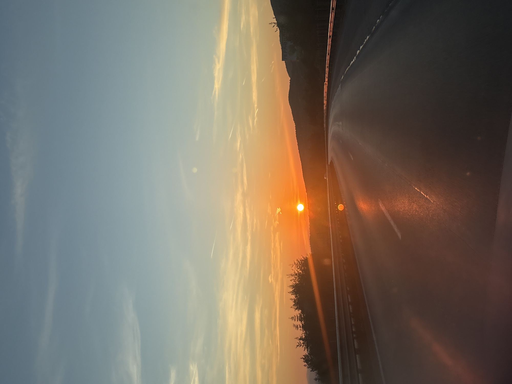 The image depicts a serene sunset view from the perspective of a vehicle traveling on a road in Stolnik, Bulgaria. The sky is dominated by the setting sun, a bright orange sphere positioned low on the horizon. Its warm light casts vibrant shades of orange, yellow, and red across the clouds and the landscape, fading into a soft blue further up.
Wispy clouds and faint contrails are visible in the sky, illuminated by the golden light. Along the horizon, a silhouette of hills or mountains stretches across the frame, appearing dark against the luminous sky, with some trees and vegetation dotting their slopes.
The foreground features a wet or highly reflective asphalt road, possibly a highway, which is intensely lit by the low sun, creating a strong glare and a broad, shimmering reflection of orange and red light. A white road marking is visible on the asphalt. To the right, a metal guardrail runs parallel to the road.
The scene captures the tranquil ambiance of late afternoon or early evening, characterized by the dramatic colors of the sunset. There are no visible people or discernible text in the image.