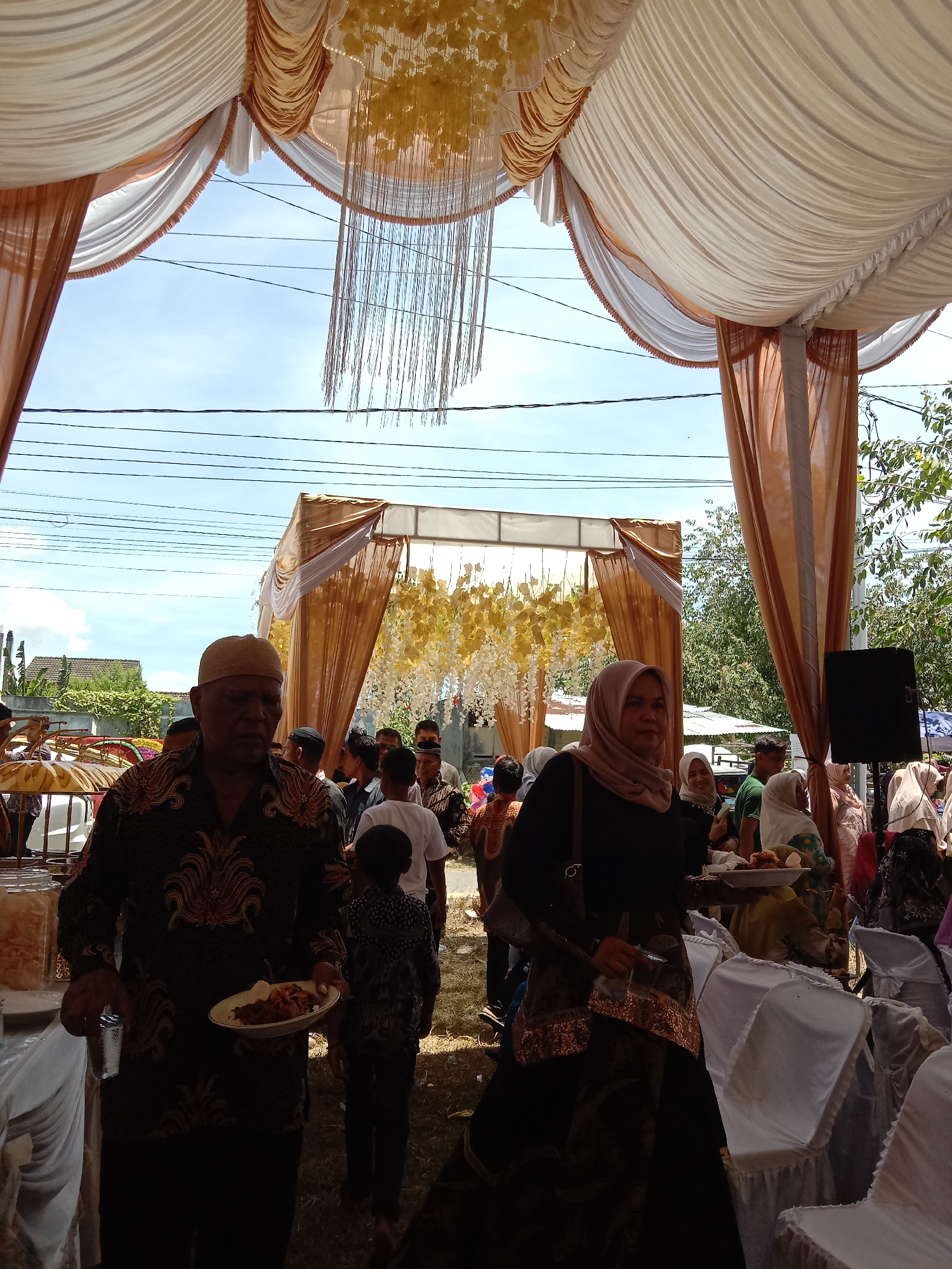 The image depicts a scene likely from a wedding reception in Lhokseumawe, Indonesia. The location appears to be outdoors, under a large white and brown tent structure with ornate floral decorations. A crowd of people is visible in the background, some interacting and others standing. In the foreground, two individuals are holding plates of food and cups. The man on the left wears a patterned shirt and a hat, while the woman on the right wears a headscarf and carries a purse. Seating with white covers is seen on the right side. Power lines are visible overhead, and general greenery and structures suggest a residential or community area.