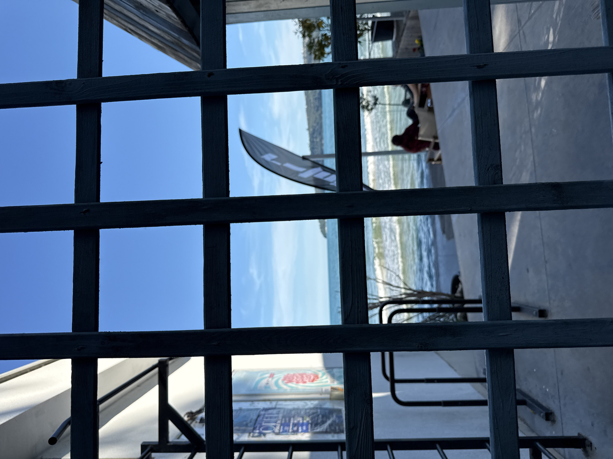 The image shows a view of the Black Sea in Varna, Bulgaria, through a dark green wooden trellis. The trellis is in the foreground and occupies a significant portion of the frame, obscuring some details of the scene beyond.
In the mid-ground, the sea is visible with small waves breaking on a sandy or pebbly shore. The water is a vibrant turquoise-blue, and the sky above is clear and bright blue, with a few wispy white clouds. A black flag with white lettering is planted on the shore.
On the shore, there are two people sitting. One person, wearing a dark red shirt, is seated in a light-colored chair, facing away from the camera. The other person's legs are visible in the foreground of the seated figures, with their feet resting on the ground.
To the left of the image, a white building with some signage is partially visible behind the trellis. The signage appears to be for a business. Also on the left, there are some black metal bicycle racks.
To the right of the image, there is a portion of a wooden structure, possibly an outdoor seating area or canopy. A small tree in a black pot is also visible to the right.
The overall scene suggests a relaxed outdoor setting by the sea, likely a cafe or a public area. The sunlight suggests it is daytime.