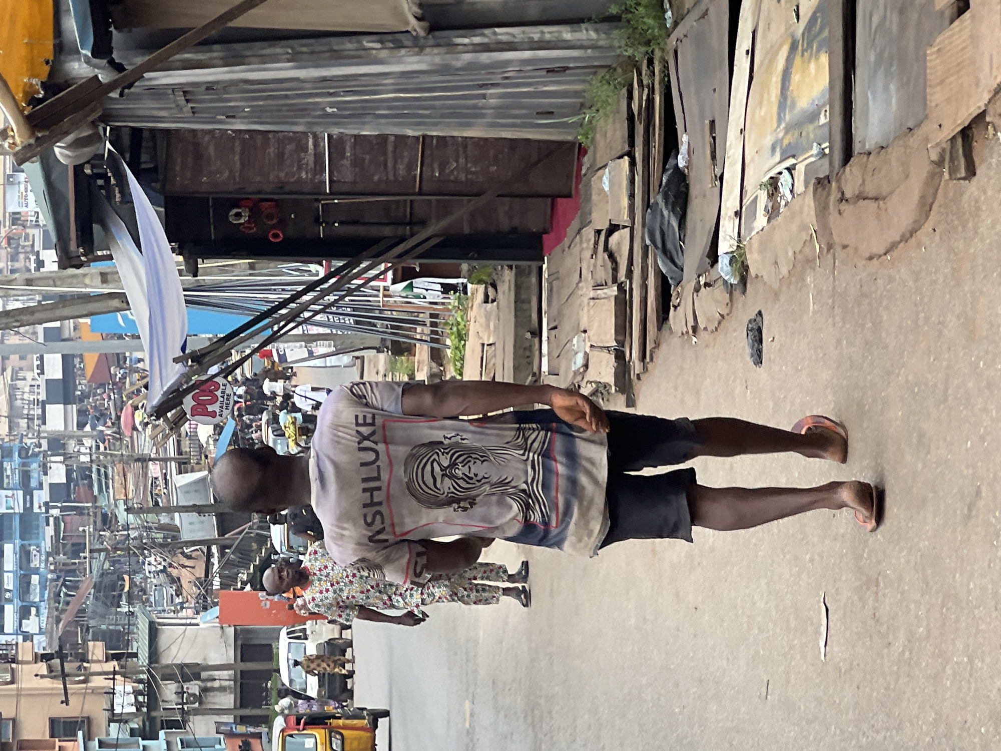 This image captures a street scene in Nkpor, Nigeria. In the foreground, a man is walking away from the camera, wearing a grey t-shirt with a graphic of a face and the word "ASHLUXE" on the back, and dark shorts. He is wearing open-toed sandals. To his left, another man in a brightly patterned shirt and trousers is walking in the same direction. Behind them, a yellow auto-rickshaw is visible, along with other vehicles and pedestrians. The background shows a densely built urban environment with numerous buildings, overhead wires, and makeshift structures. The street itself appears to be paved, with some debris and damaged sections visible along the sides. The overall atmosphere suggests a busy, commercial area.