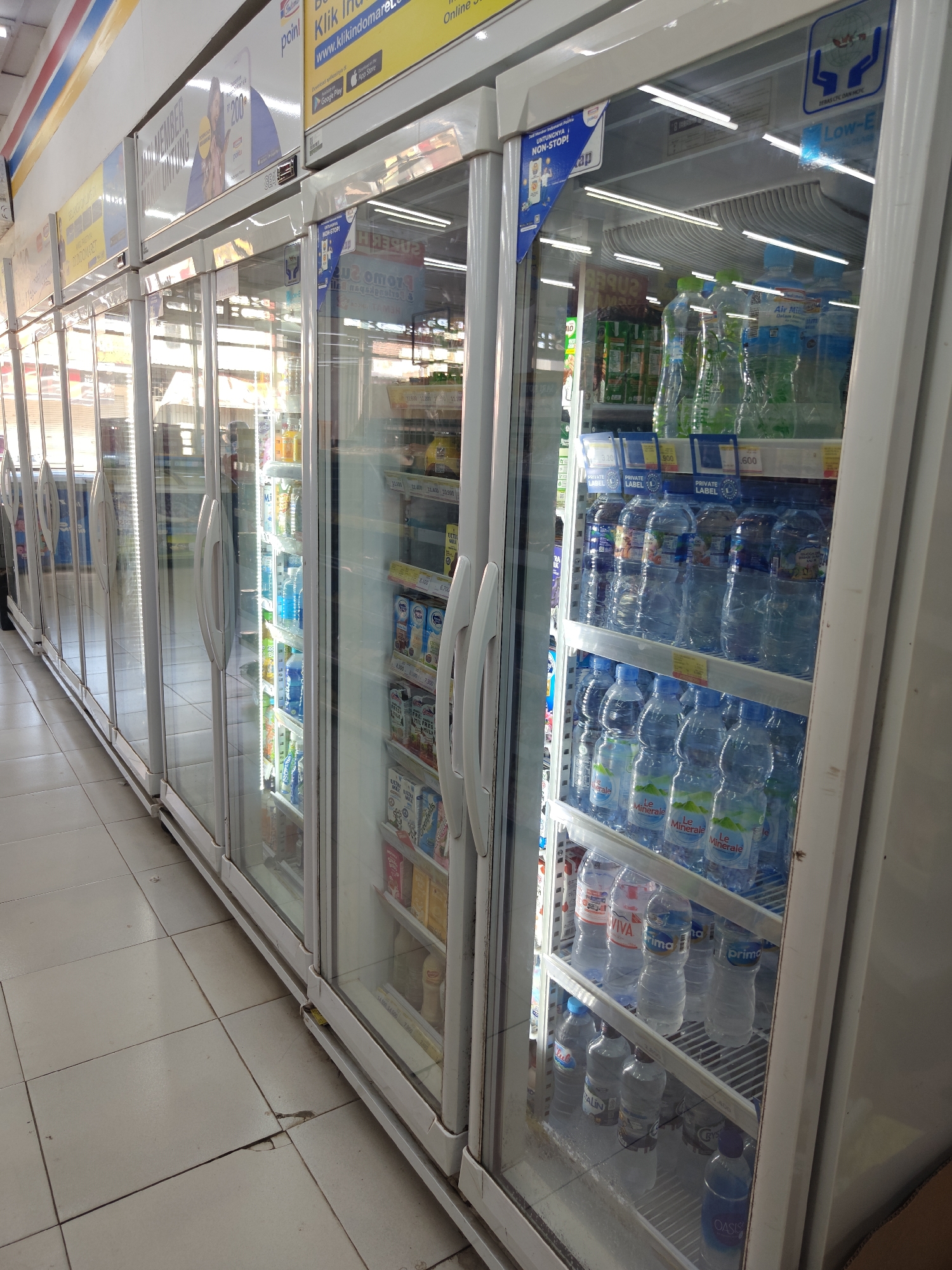 The image shows a row of commercial refrigerators with glass doors in what appears to be a convenience store, possibly in Klampokan, Indonesia. The refrigerators are stocked with various bottled beverages, primarily water. The shelves are illuminated by fluorescent lights, and some of the bottles have labels visible, such as "Le Minerale" and "Oasis". There are also some boxes of packaged drinks on the shelves. The floor is tiled and appears to be a light beige color. Above the refrigerators, there are advertising signs for a store loyalty program, displaying the "point" logo and a QR code. The overall environment suggests a retail setting designed for chilled product sales.