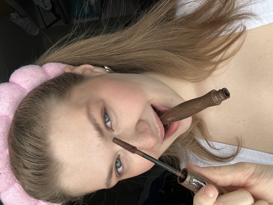 A light-skinned woman is shown from the chest up, angled downward, with a pink headband on her forehead. She holds a brown mascara tube in her hand and a mascara wand points toward her left eyebrow. A second brown mascara tube is held in her mouth. She has fair hair, bluish-gray eyes, and light eyebrows. Her lips are pink. The packaging of the mascara indicates it is "Brown Waterproof" by Catrice.