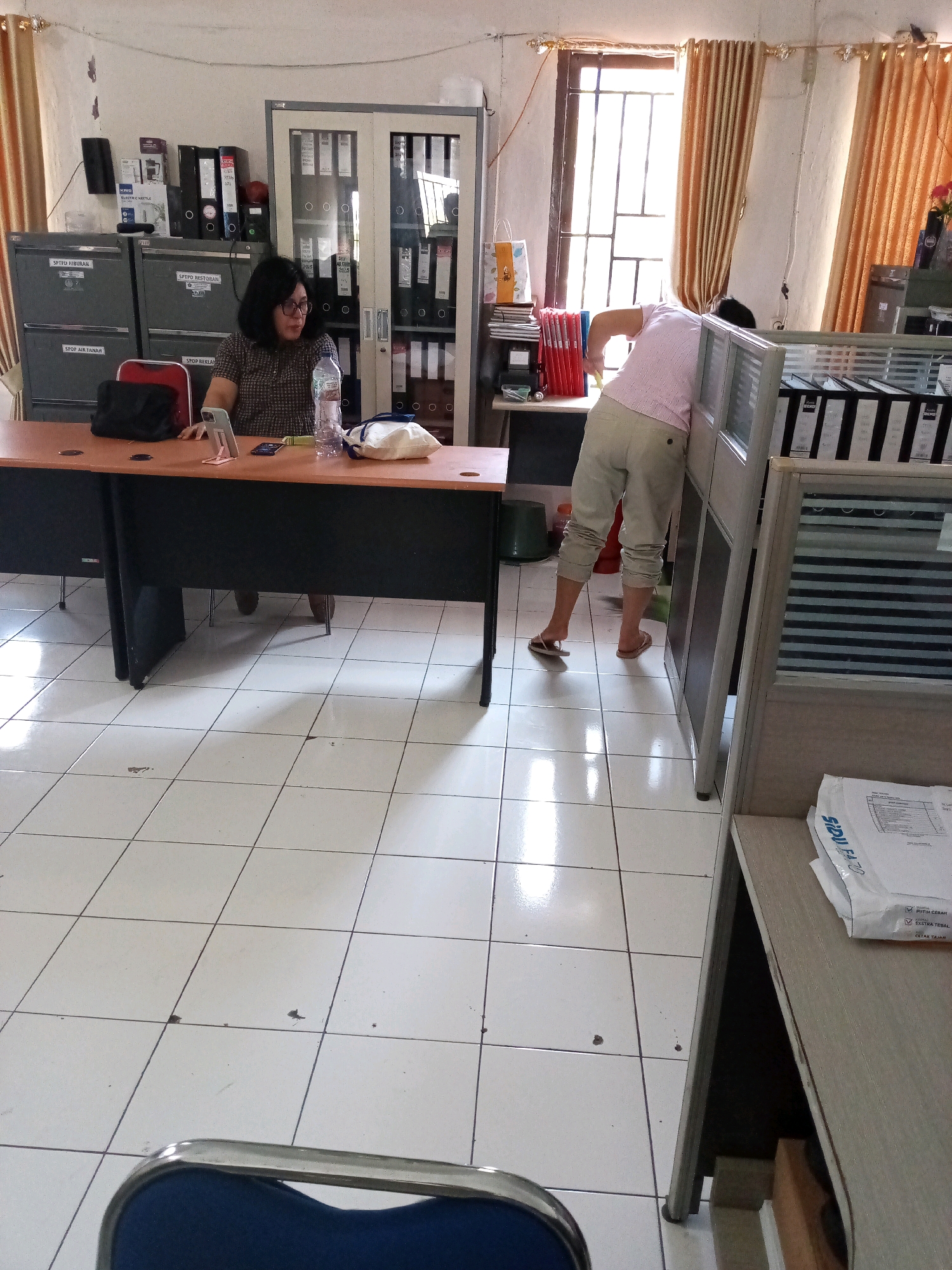 The image depicts an indoor office environment, likely a small business or government office, with white tiled flooring. In the foreground, a woman in a patterned shirt is seated at a wooden desk, looking towards her left. On the desk, there is a mobile phone in a stand, a bottle of water, and a white pillow. To the right of the desk, a person wearing a pink shirt and light-colored pants is bending over and appears to be working on something near a cubicle partition. Behind the woman, filing cabinets are visible, as well as a glass-fronted cabinet filled with binders. The room is lit by natural light streaming through orange curtains covering a window. The overall impression is one of an active workspace. The image does not provide specific clues to pinpoint the exact location in Bitung, Indonesia.