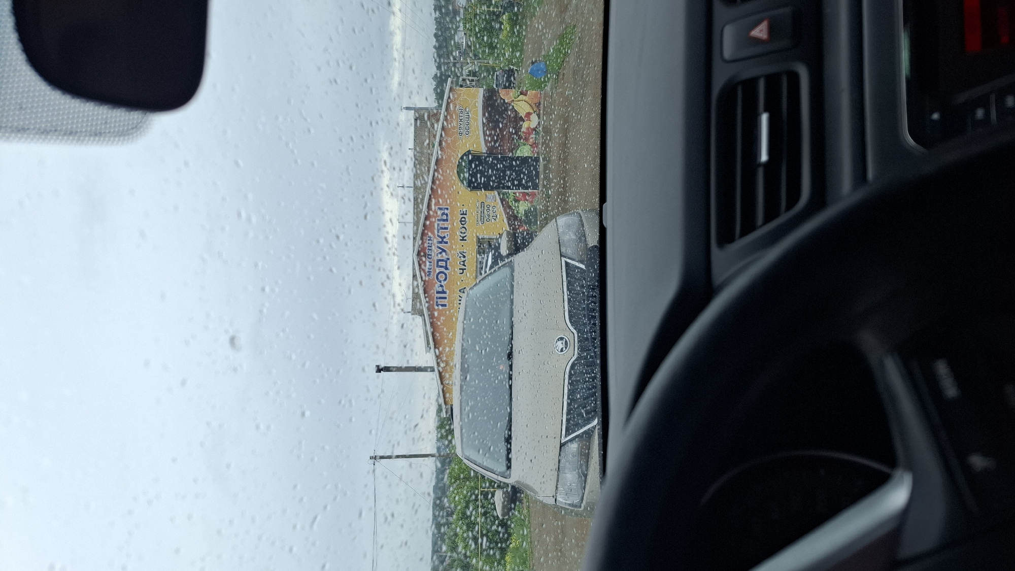 The image captures a street scene in Фоки, Russia, viewed from the interior of a car through a rain-splattered windshield. The perspective is slightly angled, looking out towards the passenger side.
Outside the car, a beige or light-colored car, possibly a Skoda indicated by its front emblem, is parked or waiting, also covered in raindrops. Behind this car is a single-story commercial building with a large, colorful mural-like sign. The sign features text in Cyrillic, identifying the establishment as a "МАГАЗИН" (Shop) selling "ПРОДУКТЫ" (Groceries/Products). Smaller text on the sign indicates items like "ЧАЙ КОФЕ" (Tea Coffee) and "ФРУКТЫ ОВОЩИ" (Fruits Vegetables), along with what appear to be operating hours, "9:00 - 20:00". The building's sign also displays various images of food products such as fruits and vegetables. Beyond the building, some greenery and utility poles are visible under a grey, overcast sky.
The weather is rainy, evident from the numerous water droplets on the car's windshield. The time of day appears to be daytime, likely overcast due to the rain.
Inside the car, parts of the dashboard are visible on the right side of the frame, including a black air vent and a red hazard light button. A section of the steering wheel is also visible in the bottom right corner, while a small part of the rearview mirror is seen in the upper left. The overall scene conveys a typical, rainy day roadside observation from a vehicle.
