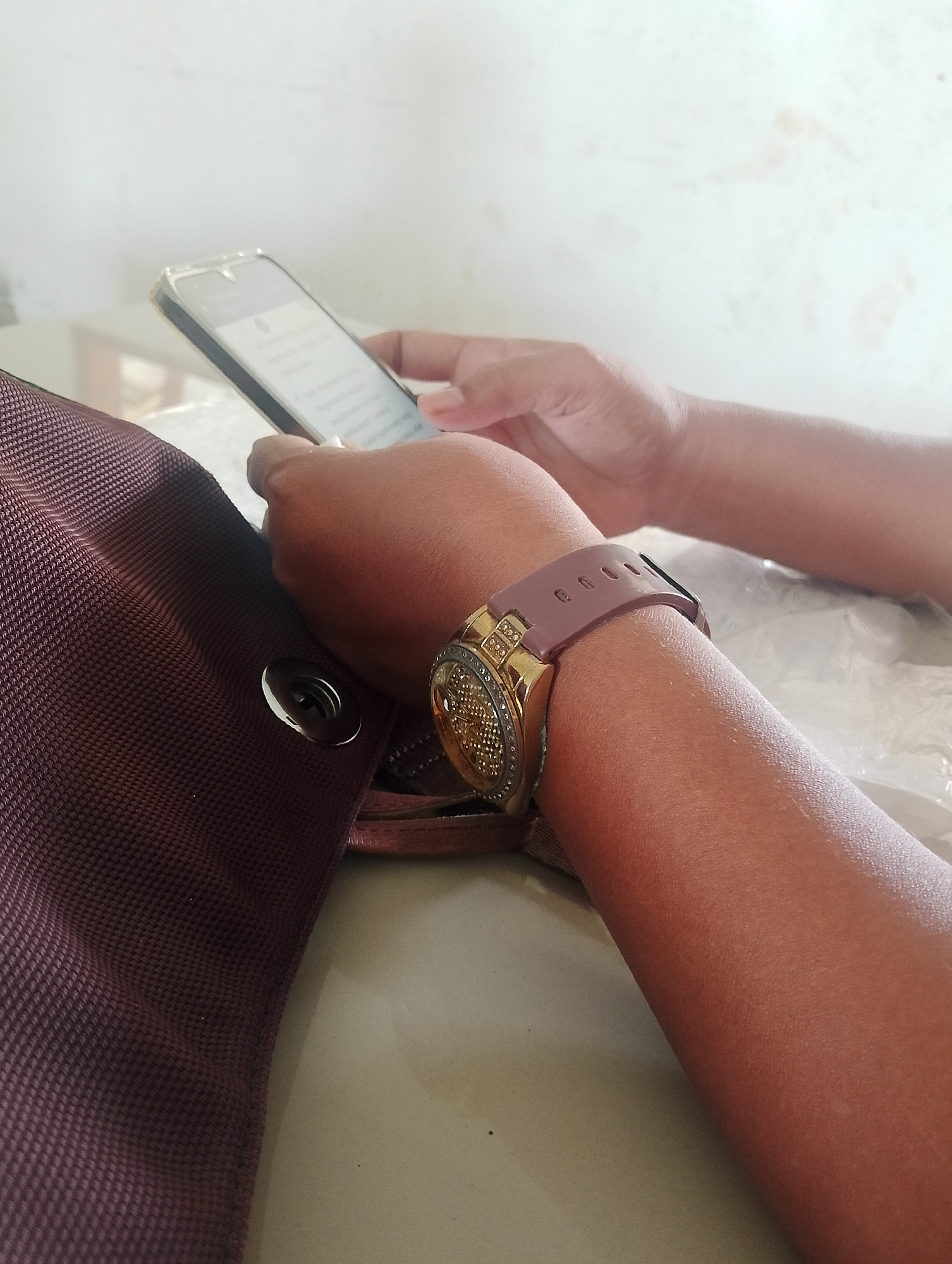 The image shows a close-up view of a person's arm and hand holding a smartphone. The person is wearing a gold watch with a pink band. The background is blurry and appears to be a wall and a table. A portion of a brown bag is visible in the foreground. The scene is likely indoors, possibly in a public place such as a cafe or waiting area, given the indistinct background. The image does not provide any specific visual cues that would allow for the localization of Serbelawan, Indonesia.