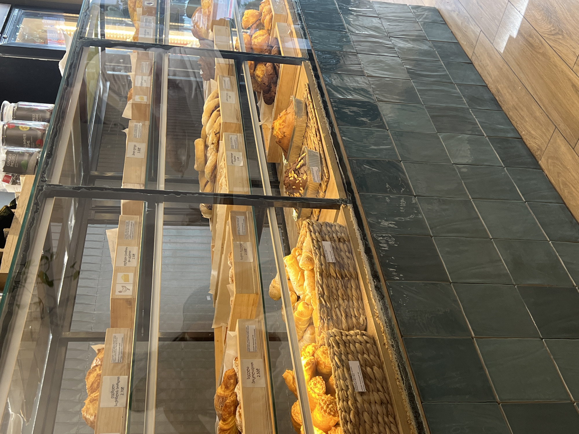 The image shows a bakery display case filled with various baked goods, including croissants, bread rolls, and muffins. The display case is made of glass and metal, and it is lit from above, highlighting the products. The baked goods are arranged in wooden trays and wicker baskets, some with price tags in Georgian currency. In the background, a refrigerator and shelves with jars of tea are visible. The overall atmosphere is clean and organized, suggesting a well-maintained establishment. The exterior of the display case is tiled with dark green, glossy tiles, and the floor is made of light-colored wooden planks. The location is a bakery in Batumi, Georgia. There are no people visible in the image.