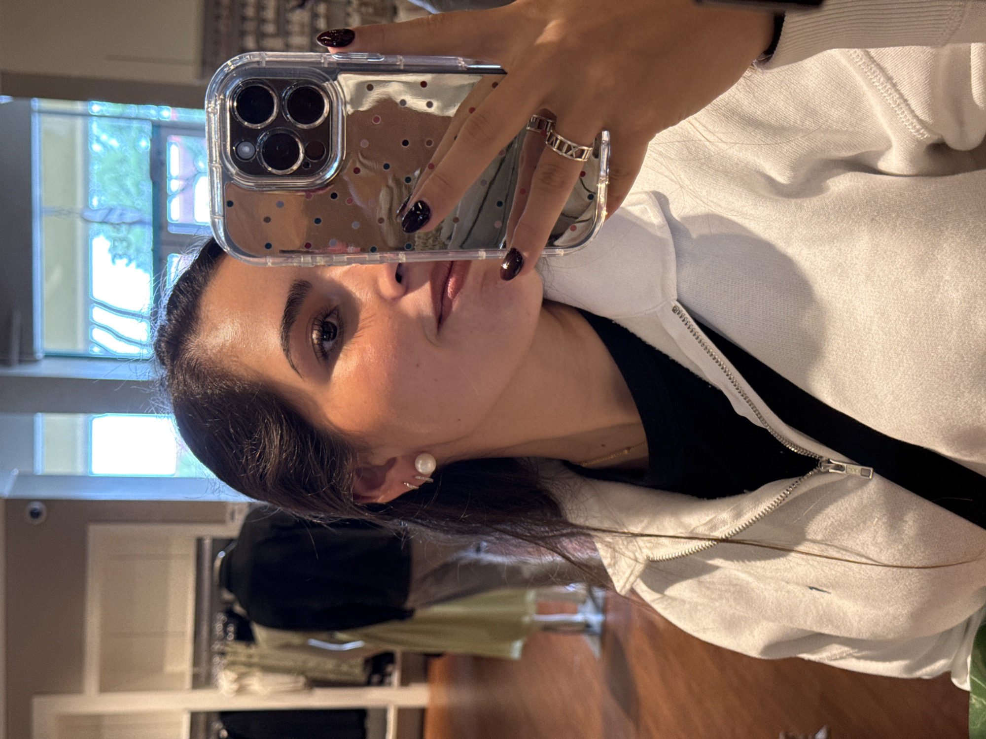 A woman takes a selfie indoors. She is holding a phone with a clear case with colored polka dots, obscuring part of her face. She has dark hair, brown eyes, dark nails, and a pearl earring. She is wearing a white zip-up jacket over a black shirt. She is also wearing rings on her fingers. In the background, there is clothing on display racks and a window with trees visible outside. The location is likely a retail store in San Marcos, Texas.