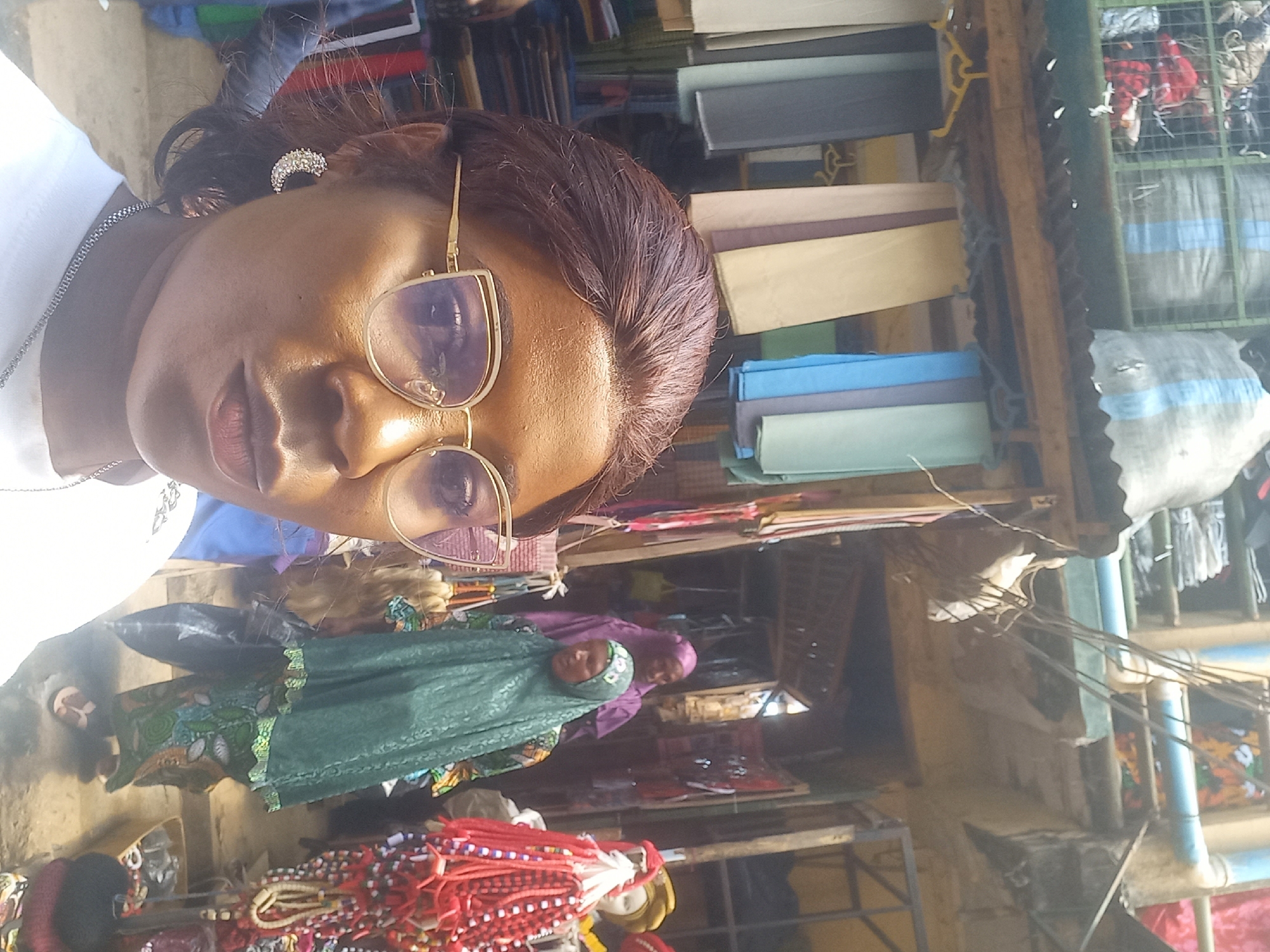 The image depicts a street market scene in Jos, Nigeria, with a woman in the foreground taking a selfie. She is wearing glasses, a white shirt, and a crescent-shaped earring. Behind her, several stalls are visible, displaying various fabrics and beaded items. Two women in hijabs are seen browsing the merchandise in the background. The market appears to be a bustling hub of commercial activity, with a variety of goods on display.