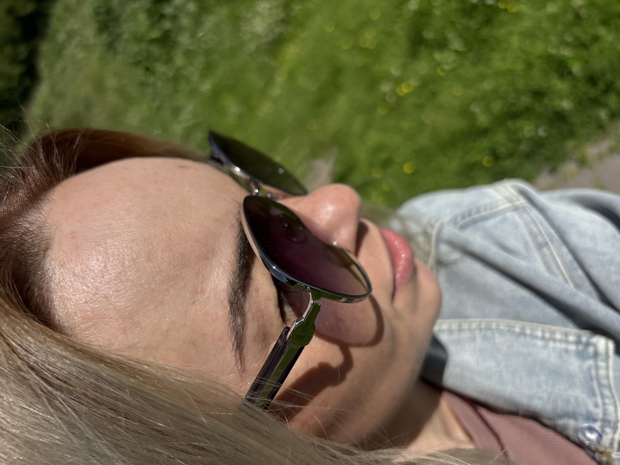 The image shows a close-up, angled shot of a young Caucasian woman with blonde hair, wearing sunglasses and a denim jacket. She is outdoors, with a blurred green background suggesting a park or grassy area with some yellow flowers. The lighting indicates it is daytime, likely sunny, casting shadows on her face. No specific landmarks of Saint Petersburg are visible.