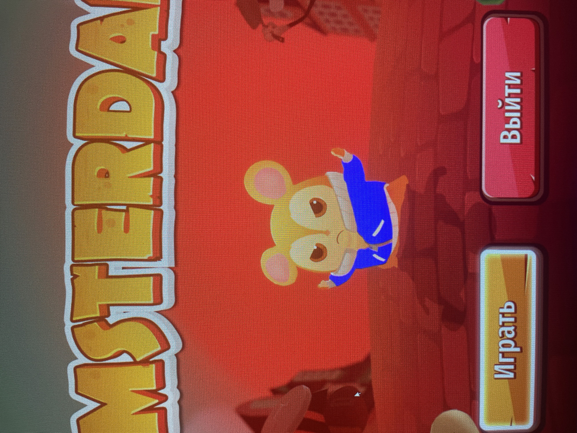 The image is a screenshot of a video game's title screen. The title, "MSTERDAM", is displayed in large, stylized yellow letters with a red outline against a gradient background that transitions from grey to green at the top and red at the bottom. In the center of the screen, a cartoonish hamster wearing a blue jacket stands with its arms outstretched. To the left and right of the hamster are blurry, dark silhouettes of buildings or structures. Below the hamster are two buttons. The left button is yellow and contains the Russian text "Играть" (Play). The right button is red and contains the Russian text "Выйти" (Exit). There are no identifiable landmarks or elements that connect this image to Sinyavino, Russia.