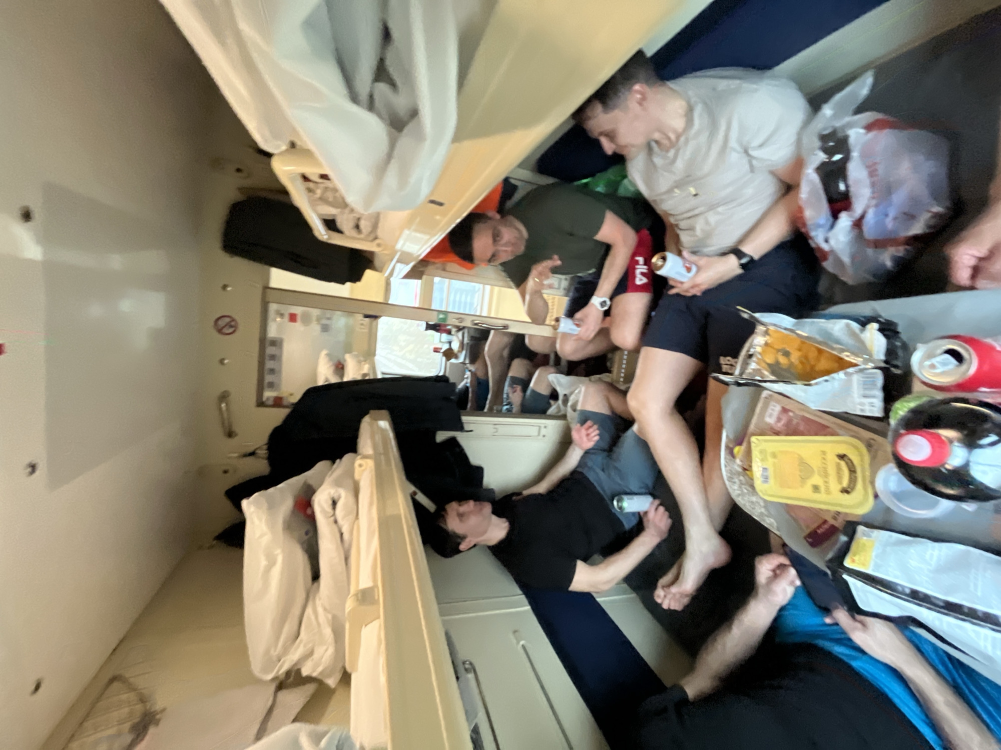 Inside a train compartment, four men are visible. One man is lying down on the lower bunk while another man sits nearby. Across the compartment, a third man sits on the upper bunk while a fourth man sits next to him. Various food items, including cans, a package of butter, and a bag, are on a table in the compartment. Clothing hangs from the upper bunk. A window is visible on the left, showing the reflection of the people in the compartment. Based on the image's prompt information, this scene is located in a train compartment, possibly traveling through or originating from Izhevsk, Russia.