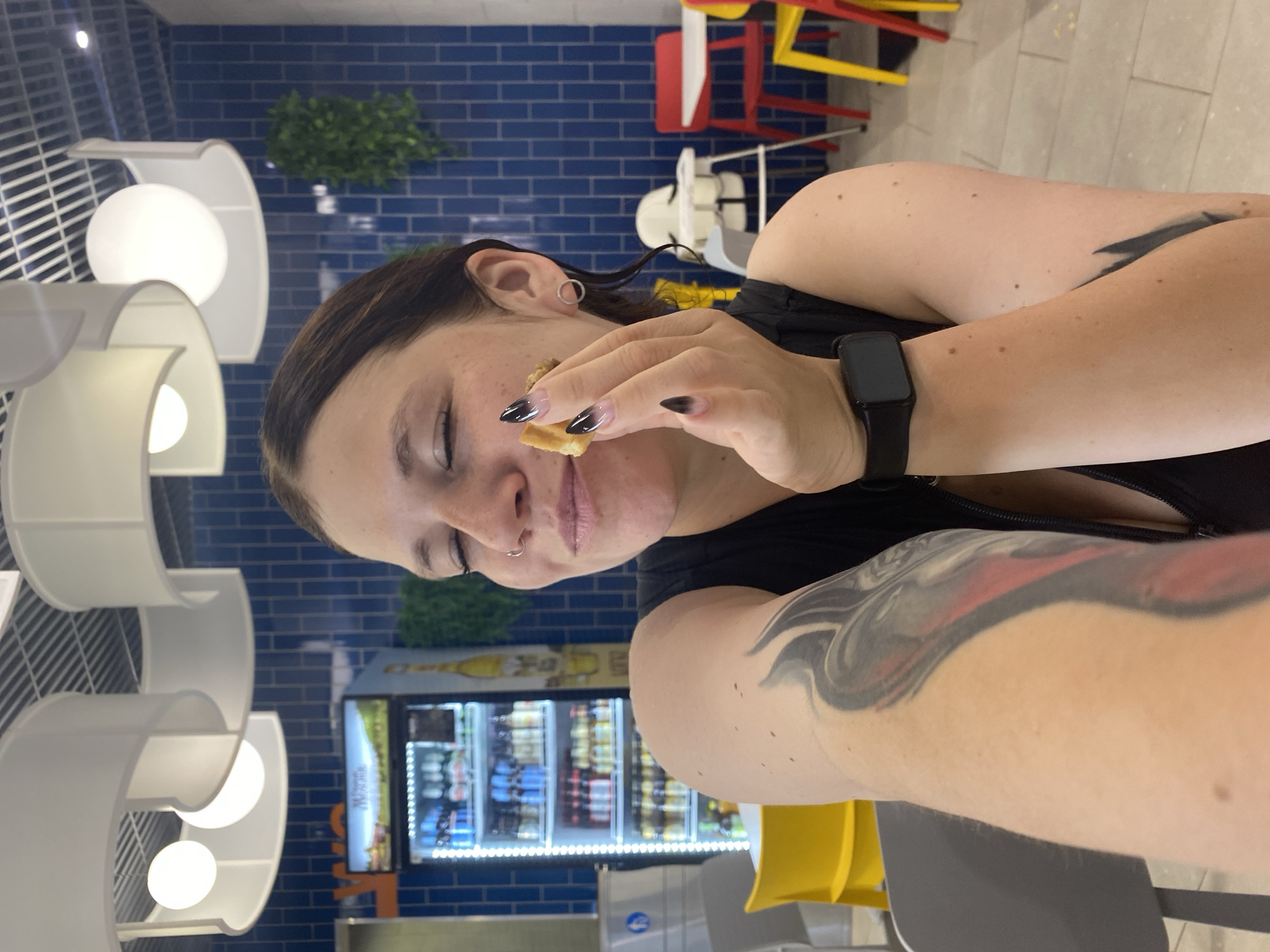 A woman is shown in a close-up shot, holding a small piece of food to her mouth. Her eyes are closed, and she appears to be enjoying the food. She has a nose ring and a tattoo on her arm. The background shows a brightly lit refrigerator filled with drinks, a blue tiled wall, and several chairs and tables. There are also modern light fixtures on the ceiling. The scene appears to be in a cafe or restaurant. There is no information available to confirm the location in Votkinsk, Russia.
