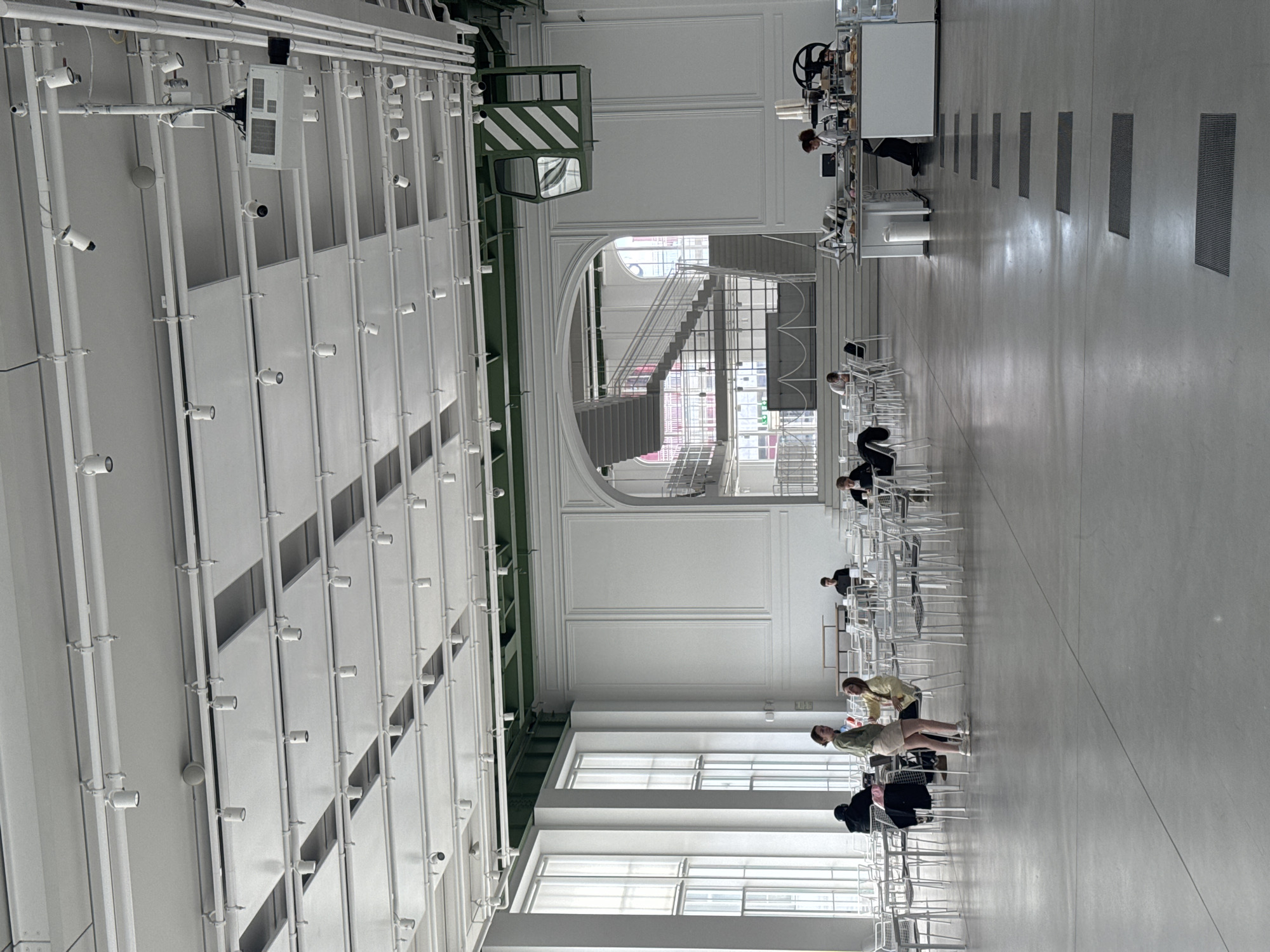 This media file captures the interior of a spacious, modern hall in Moscow, Russia, characterized by high ceilings and a minimalist design, with the image oriented vertically to show the room's length.
The ceiling is a notable feature, adorned with numerous exposed white pipes, track lighting fixtures, and air ducts, creating an industrial-chic aesthetic. A prominent, dark green overhead structure with diagonal white stripes, possibly an industrial crane or beam, traverses the upper left portion of the room. The walls are predominantly white, featuring large, rectangular windows along the lower half of the right wall (when viewed upright), which allow ample natural light to illuminate the space.
Several people are present in the mid-ground and background. Multiple individuals are seated at white tables with matching white chairs, some appearing engaged in work or quiet conversation. In the far background, a long white counter serves as a service area, with at least one person working behind it, surrounded by coffee machines and cups. A large, curving staircase with metal railings ascends in the background, adding a dynamic architectural element. The floor is a light grey, reflective surface, interspersed with several rectangular floor grates.
In the foreground, two women stand near a row of white chairs, one wearing a light yellow top and white shorts, and the other a light green top and light-colored shorts; they appear to be interacting or pausing. The overall atmosphere is bright and airy, suggesting it is daytime. No visible text is legible within the image.
