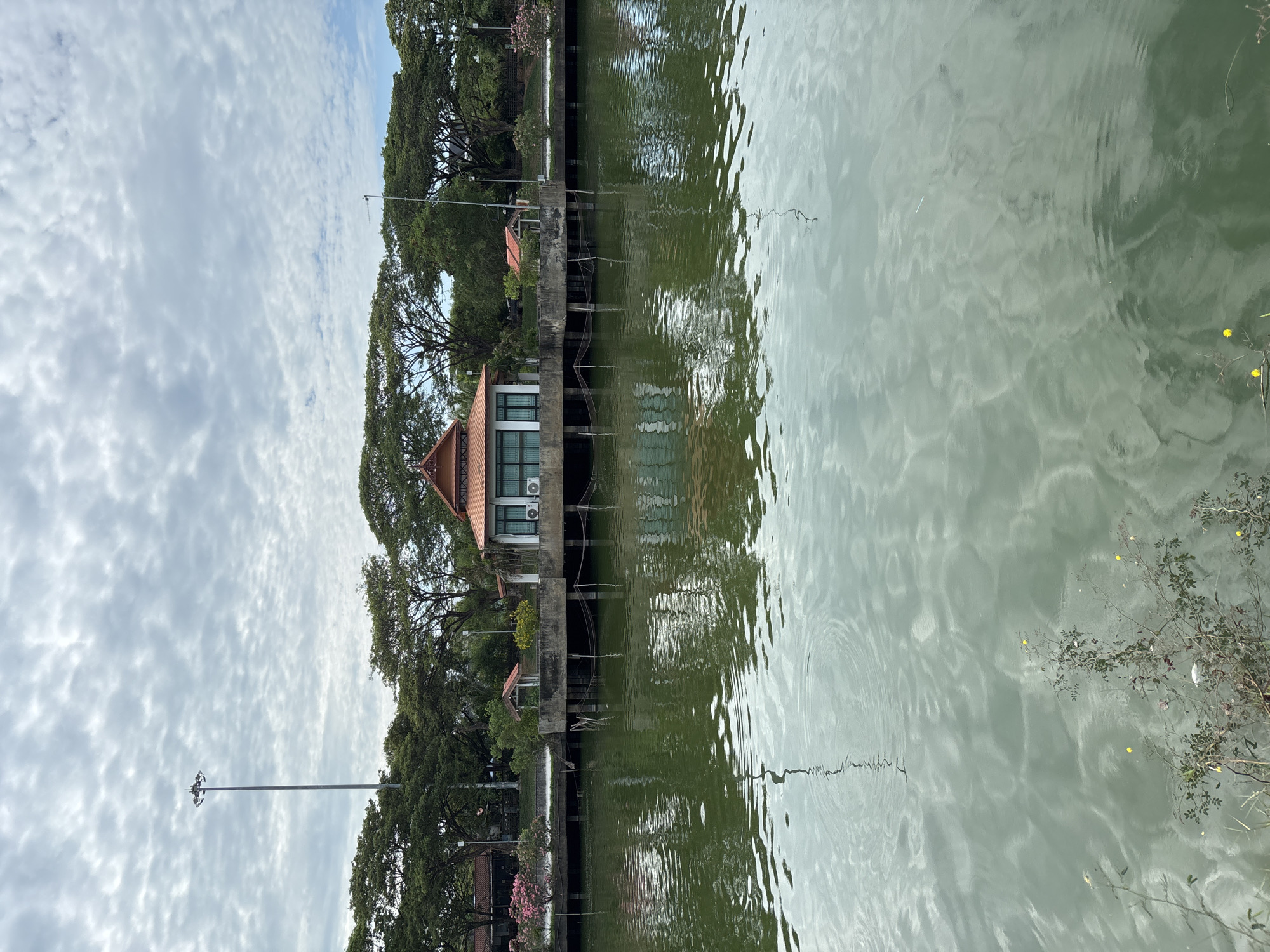 The image shows a body of water, likely a lake or pond, with a building constructed on stilts extending into the water. The architecture of the building suggests a Thai style. Lush green trees line the bank of the water. The sky is overcast with layered cloud formations. Reflections of the trees and buildings are visible in the water. Some vegetation, including flowering plants, are along the edges of the water. The location is Baan Chaweng, Thailand.