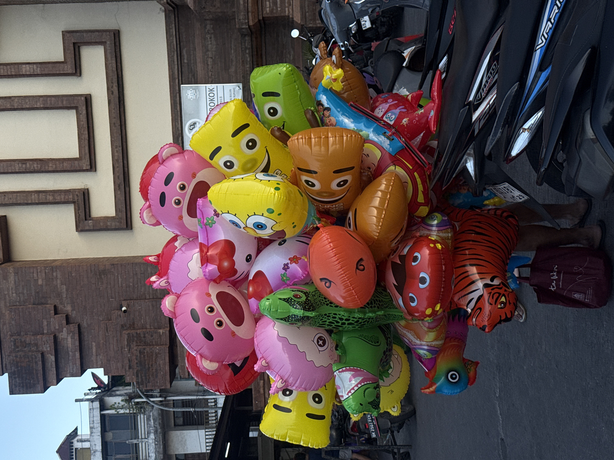 The image depicts a close-up view of a large assortment of colorful balloons, predominantly in character shapes. The balloons are clustered together, with several visible characters including pink bear-like faces, a yellow character with wide eyes and a smile, a green dinosaur-like character, a tiger, and an orange fish-like character. There are also other brightly colored balloons with various designs. The balloons are held by an unseen person, with only their legs and feet visible at the bottom right of the frame. A dark red reusable bag rests on the ground near their feet. In the background, several motorbikes are parked. Behind the motorbikes, there is a building with brown brickwork and a light-colored facade with decorative elements. A sign is visible on the building, though its text is partially obscured. The scene appears to be on a street in Denpasar, Indonesia, as indicated by the presence of motorbikes and typical urban architecture. The ground is paved with asphalt. The lighting suggests it is daytime, possibly late afternoon given the shadows.