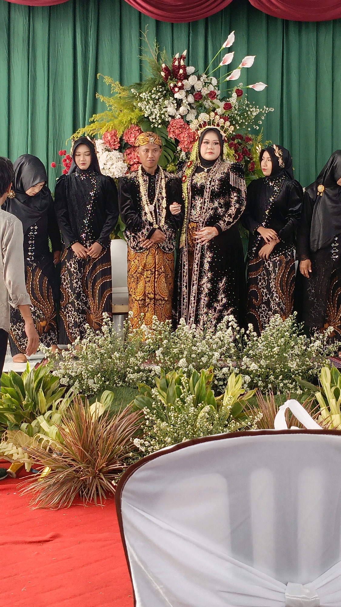 The image depicts a wedding ceremony in Gondek, Indonesia, with a bride and groom standing at the center. They are dressed in traditional attire. The groom wears a patterned sarong, a dark velvet top adorned with intricate gold embroidery, and a gold headpiece. The bride is in a black dress with elaborate gold embellishments and a matching headpiece, jewelry, and makeup. They are flanked by several women wearing black hijabs and matching patterned skirts. In the background, a large floral arrangement with red, white, and pink flowers is displayed against a green curtain. The foreground features lush greenery and a red carpet. A white cloth-covered chair is visible on the right side of the image.
