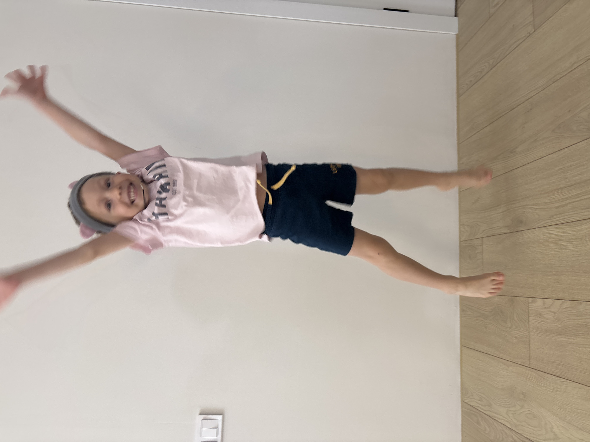 A child is positioned horizontally in the center of the image, appearing to float against a white wall and a light brown, wood-patterned floor. The child is wearing a pink t-shirt, dark blue shorts, and a gray headband with what appear to be small ears attached. Both arms are raised, with the hands open. The child is smiling. The child is barefoot. A light switch is visible on the wall, near the bottom of the image. It is not possible to determine the location or city from this image.