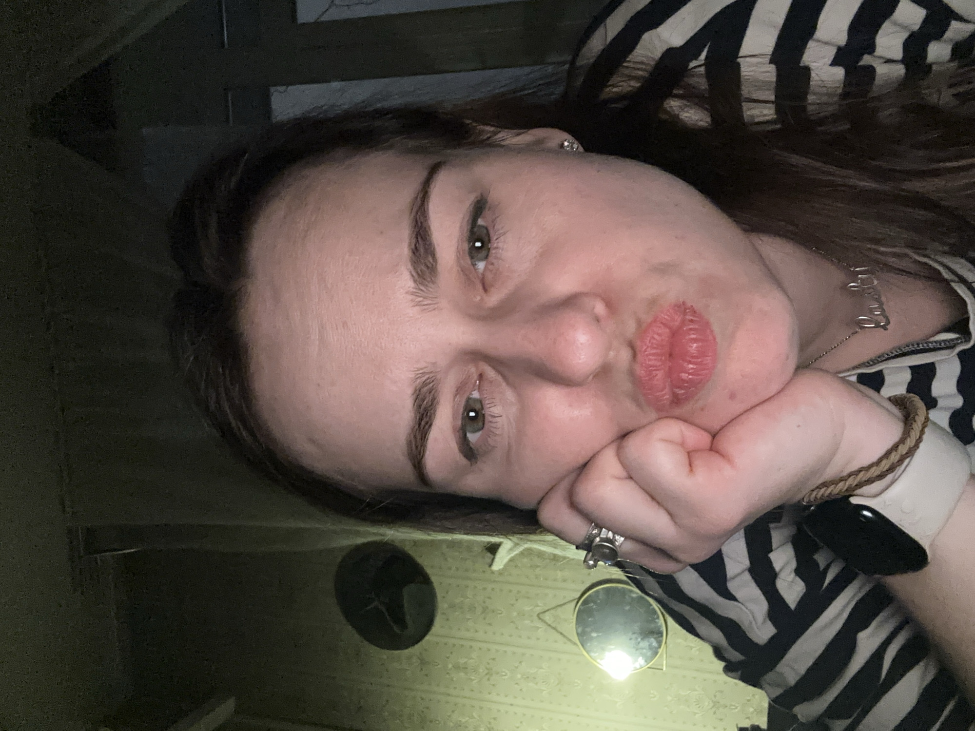 A woman with fair skin, brown hair, and green eyes poses in a selfie. She rests her face on her hand, displaying rings on her fingers, a white smart watch with a black screen, and a braided bracelet. She wears a striped black and white shirt and a necklace with a word. The background includes a textured wall with a round mirror and a dark object, possibly a hat. A staircase is visible in the top left corner. There is no information indicating the location is Moscow, Russia.