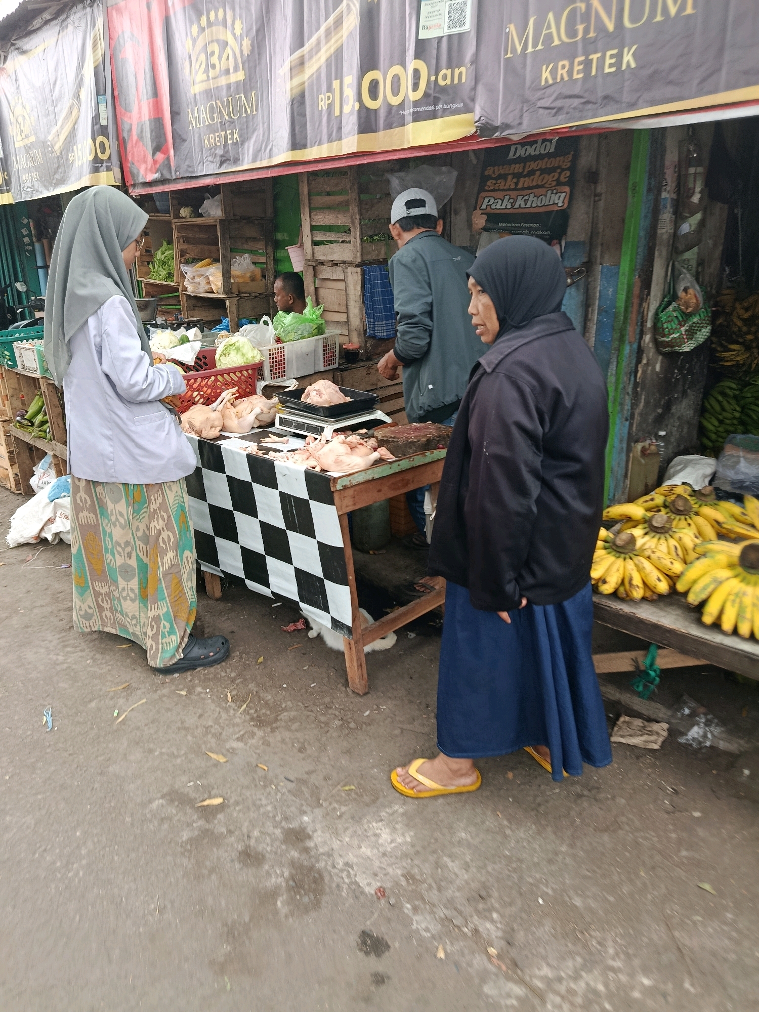 In Bululawang, Indonesia, a street market scene unfolds with three individuals engaged in commerce. A woman wearing a grey hijab and a light-colored jacket stands at a market stall, her gaze directed downwards towards the goods. Beside her, a man in a grey jacket and a white cap appears to be a vendor, his hands resting on the counter. Another woman, draped in a black hijab and a dark jacket, stands to the right, observing the scene.
The market stall is adorned with a black and white checkered tablecloth, displaying raw chickens and other produce. Behind the stall, wooden crates are stacked with various items, including vegetables and bags of what appears to be food. The backdrop features a large banner with text in Indonesian, promoting products like "MAGNUM KRETEK" and advertising prices. To the right of the stall, a large bunch of yellow bananas is prominently displayed on a wooden surface. The ground beneath is paved with asphalt, showing signs of wear and scattered debris.