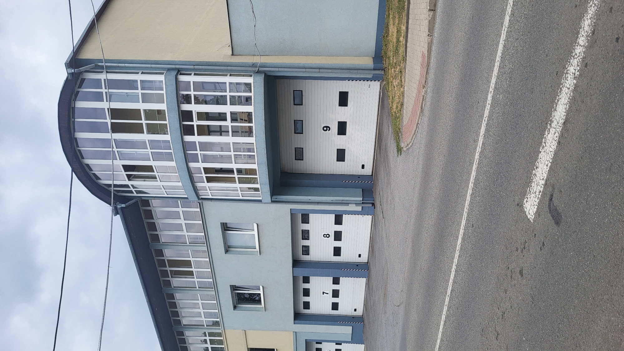 The image shows the exterior of a fire station in Prešov, Slovakia. The building is light blue with a beige section on the right. It features a curved glass facade on the upper floors and three visible garage doors on the ground level, numbered 7, 8, and 9. The garage doors are white with dark square windows. In front of the building is a paved road with white lane markings. A grassy area with a brick border is visible to the right of the building. The sky is overcast.