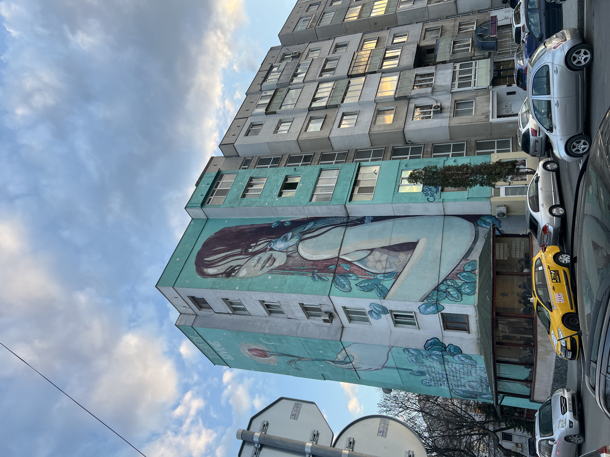 The image depicts a multi-story residential building in Sofia, Bulgaria, prominent for a large mural covering two of its facades. The mural, rendered in teal, light blue, and white tones, portrays a stylized woman with long dark hair, a small blue bird on her right shoulder, and her left hand holding a red rose on a long stem. Stylized foliage elements are incorporated around the lower sections of the artwork. Vertical text "THINKING OF GIVING" is partially visible on the left facade.
The building itself is a block of flats with multiple windows. Adjacent to the mural building on the right are similar multi-story residential structures, characterized by grey and off-white concrete facades with numerous windows, some reflecting warm, possibly late afternoon, sunlight.
At street level, the base of the mural building features a single-story glass-enclosed structure, likely a cafe or restaurant, where several individuals are seated inside. The street in the foreground is lined with parked cars, including a yellow Renault station wagon taxi displaying Cyrillic "ТАКСИ" and a phone number (912 63), confirming the Bulgarian context. A silver Kia Carens with a "CH" license plate prefix is visible near the taxi.
Further to the left, a metal pole with the backs of two circular street signs is present. Bare deciduous trees are visible on the left, and a slender evergreen tree stands near the mural's base. The sky above is partly cloudy with patches of blue and white.