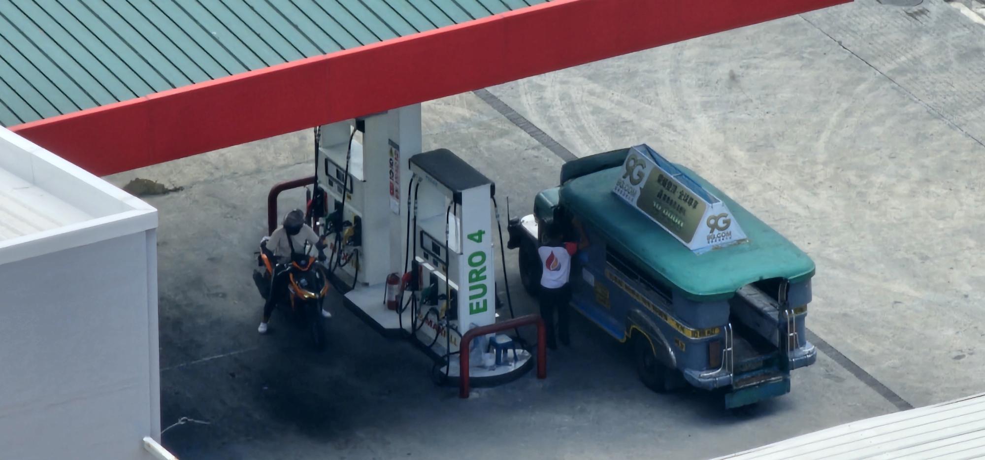 The image shows a view from above of a gas station canopy in Makati, Philippines. A green and blue jeepney is parked at a pump, with a person in a white shirt with a flame logo filling it. A person on a motorcycle with a black helmet is at another pump, also seemingly filling up. The gas station has white and red structures, with a green metal roof. The ground is concrete. A white building is visible in the foreground to the left. The scene appears to be during daylight.