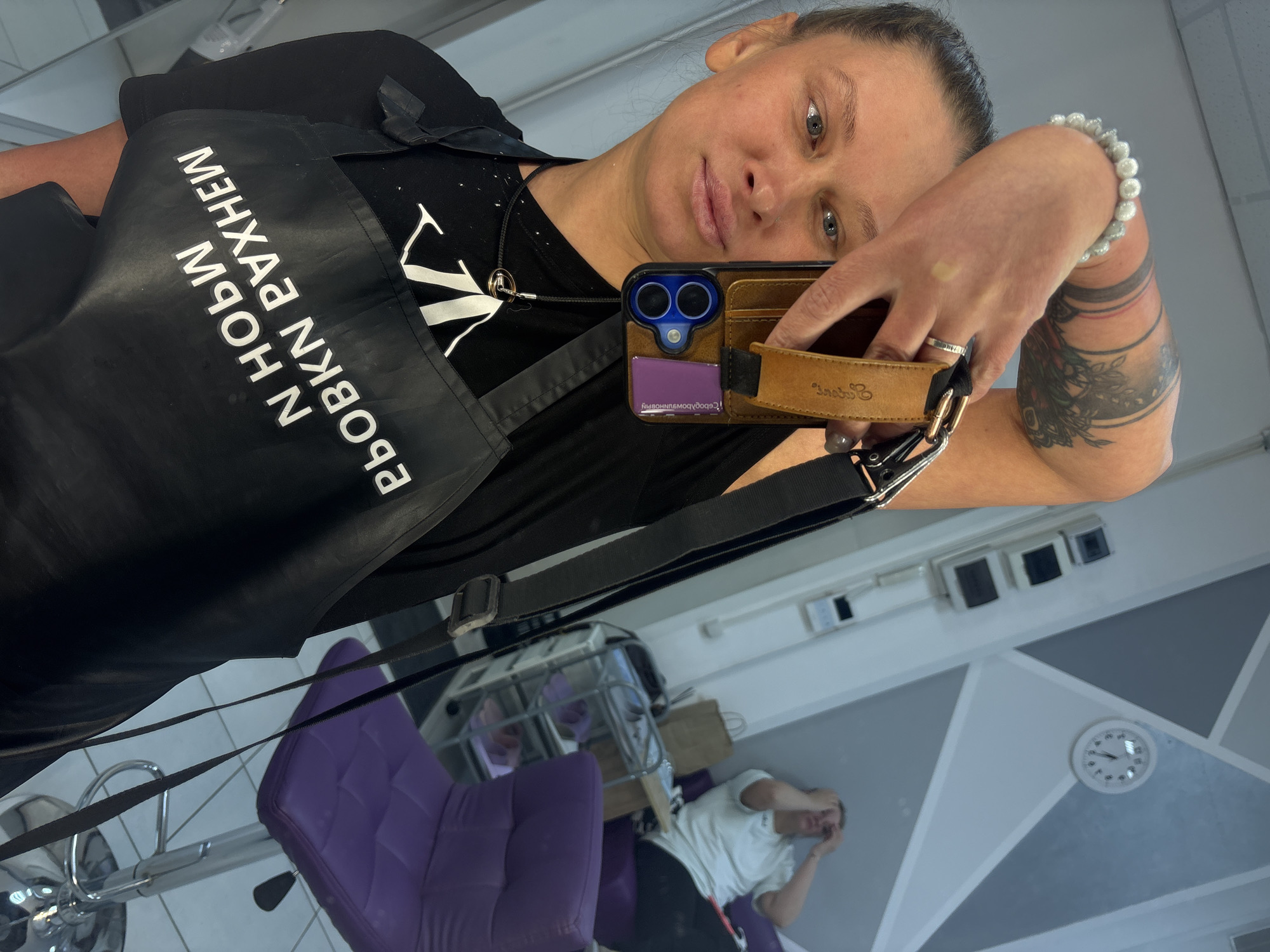 The image is a mirror selfie taken indoors in what appears to be a salon or professional studio in Perm, Russia. The main subject is a woman taking the selfie, looking directly into the camera/mirror. She has light eyes, her hair is pulled back, and she has a neutral expression. She wears a black t-shirt and a black apron with white Cyrillic text, which appears mirrored and partially upside down due to the reflection. The visible text on the apron reads "МЭНХАА РУОВКН N ЬОН Н".
Her left arm, adorned with a white beaded bracelet and silver rings, holds a dark-colored smartphone with a prominent dual-camera setup within a brown, leather-like wallet case. A purple card is visible in the phone case. A black strap or lanyard is attached to the phone case and extends around her neck. Her forearm displays a black, detailed tattoo with floral or leaf-like patterns and abstract lines.
In the reflection of the mirror, behind the woman, another person is visible, sitting in a purple, tufted styling chair. This person is wearing a white t-shirt and has their hands over their head or face. The room features a light-colored tiled floor and walls with geometric patterns in shades of gray and white. A white round wall clock shows the time to be approximately 10:15 or 10:20. The lighting is bright, suggesting daytime.