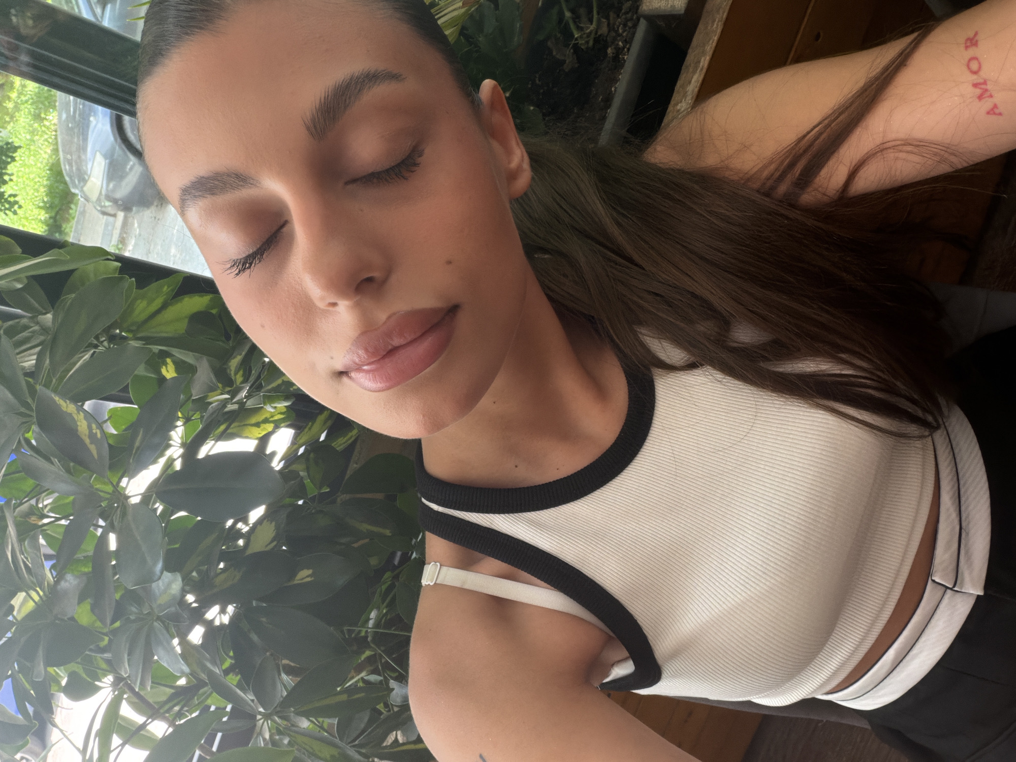 This is a close-up, portrait-style selfie of a young woman. She has her eyes closed and is looking slightly upwards, with a soft, relaxed expression. Her hair is dark brown and pulled back into a ponytail. She is wearing a white ribbed tank top with black trim. A tattoo is visible on her upper arm, spelling out "AMOR" in red lettering. The background is slightly out of focus, showing greenery, suggesting an outdoor or indoor setting with plants. The lighting appears natural, possibly indicating daytime. The image has a personal, intimate feel. The location context of Burgas, Bulgaria, is noted but not visually evident in the image itself.