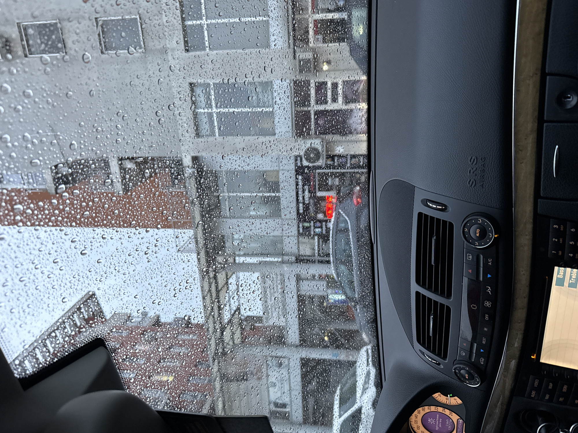 The image depicts a rainy day scene viewed from inside a car. Raindrops heavily cover the windshield, partially obscuring the view of the outside environment. Through the rain-streaked glass, multi-story buildings are visible, suggesting an urban setting. The architecture includes a mix of styles and construction materials, with white and red brick buildings prominent. Some buildings show signs of aging and weather exposure. Parked vehicles line the street.
The interior of the car features a dashboard with visible controls, including those for air conditioning and other in-car systems. The dashboard materials include wood trim. The car's speedometer is visible, indicating a speed of 0 km/h. A screen displays information, including settings for "Bass" and "Treble" controls.
Based on the license plate lamp warning on the dashboard, the location could be in a European country. Given the architectural style of the buildings, it is possible that the image was taken in Sofia, Bulgaria.