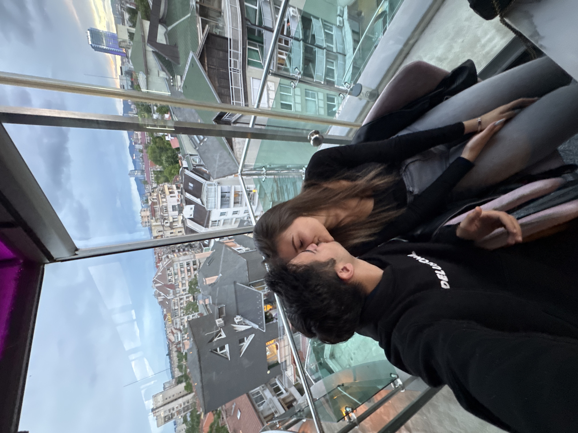 A medium shot captures a kissing couple inside a high-rise building, with the cityscape of Sofia, Bulgaria, visible through the glass windows behind them. The woman has long brown hair and wears a long-sleeved black top. The man is close to her, wearing a black sweatshirt. They are seated together. The background shows Sofia’s urban landscape, including buildings, rooftops, and a view of the sky. Reflections on the glass create a layered effect, merging the interior and exterior scenes.