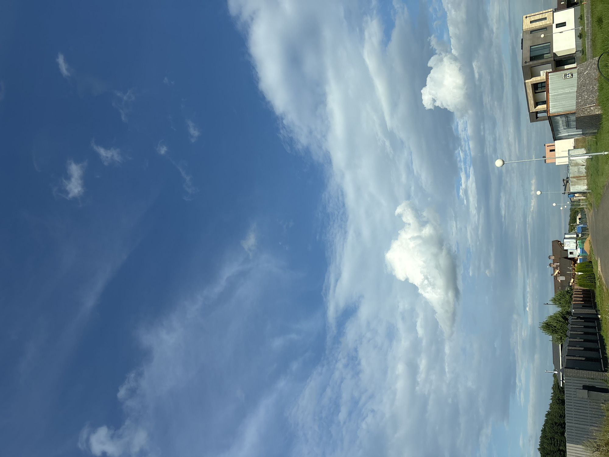The image depicts a suburban or rural street scene under a dynamic sky. The sky is a prominent feature, with a deep blue hue transitioning into lighter shades towards the horizon, and adorned with scattered white clouds, some wispy and others more substantial. A road runs through the center of the frame, leading into the distance.
On either side of the road, various houses and buildings are visible. To the left, there is a low fence made of dark bricks and grey concrete or stone panels, with some greenery behind it. Further back, a dense line of dark green trees forms a backdrop. Several houses with pitched roofs in brown and grey tones are also present on the left.
On the right side of the road, modern architectural styles are evident. One house is particularly striking with its geometric design, featuring a combination of light-colored walls, dark grey facade elements, and large windows. Adjacent to it is a corrugated metal structure. Several white streetlights with spherical lamps are spaced along the road.
The environment appears to be a developing residential area, possibly on the outskirts of a town or city, given the mix of existing structures and more contemporary buildings. The overall impression is one of a bright, possibly late afternoon or early morning, with a sense of open space and a mix of natural and constructed elements. There are no people visible in the image.