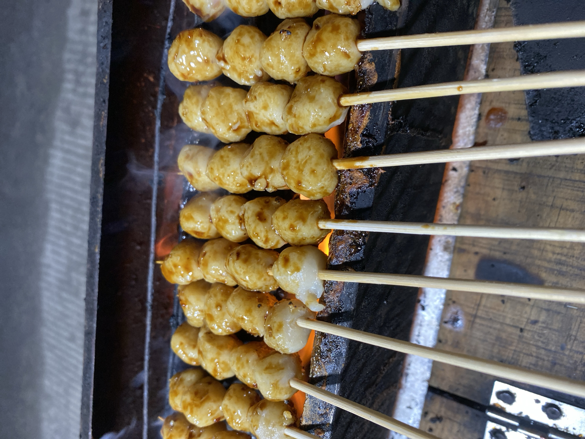 The image shows rows of grilled meatballs on skewers over a charcoal grill. The meatballs are small, round, and coated in a glossy, brownish sauce. They are arranged in multiple rows, with each row containing several skewers. The skewers are made of wood and are inserted into the meatballs. The grill appears to be made of dark metal, with glowing embers visible beneath the meatballs, indicating heat and cooking. The background shows the edge of the grill and a wooden surface below it. The lighting suggests it might be evening or a dimly lit outdoor setting. This scene is likely from a street food vendor in Kasihan, Indonesia, where grilled meatballs are a popular snack.