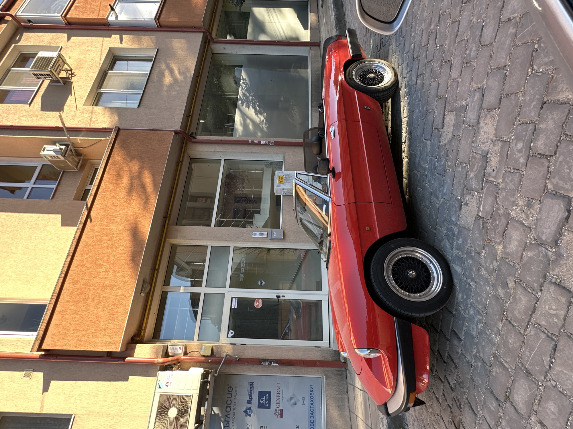 A red convertible sports car is parked on a cobblestone street in front of a building. The building has multiple windows and air conditioning units mounted on its exterior. Signs with text in Cyrillic are visible on the building's lower level, including logos for "Армеец", "UNIQA", and "GENERALI". The car has black rims and is parked parallel to the building. The sun is casting shadows, suggesting it is daytime. The scene appears to be a street in a town or city, possibly in Bulgaria given the Cyrillic text.