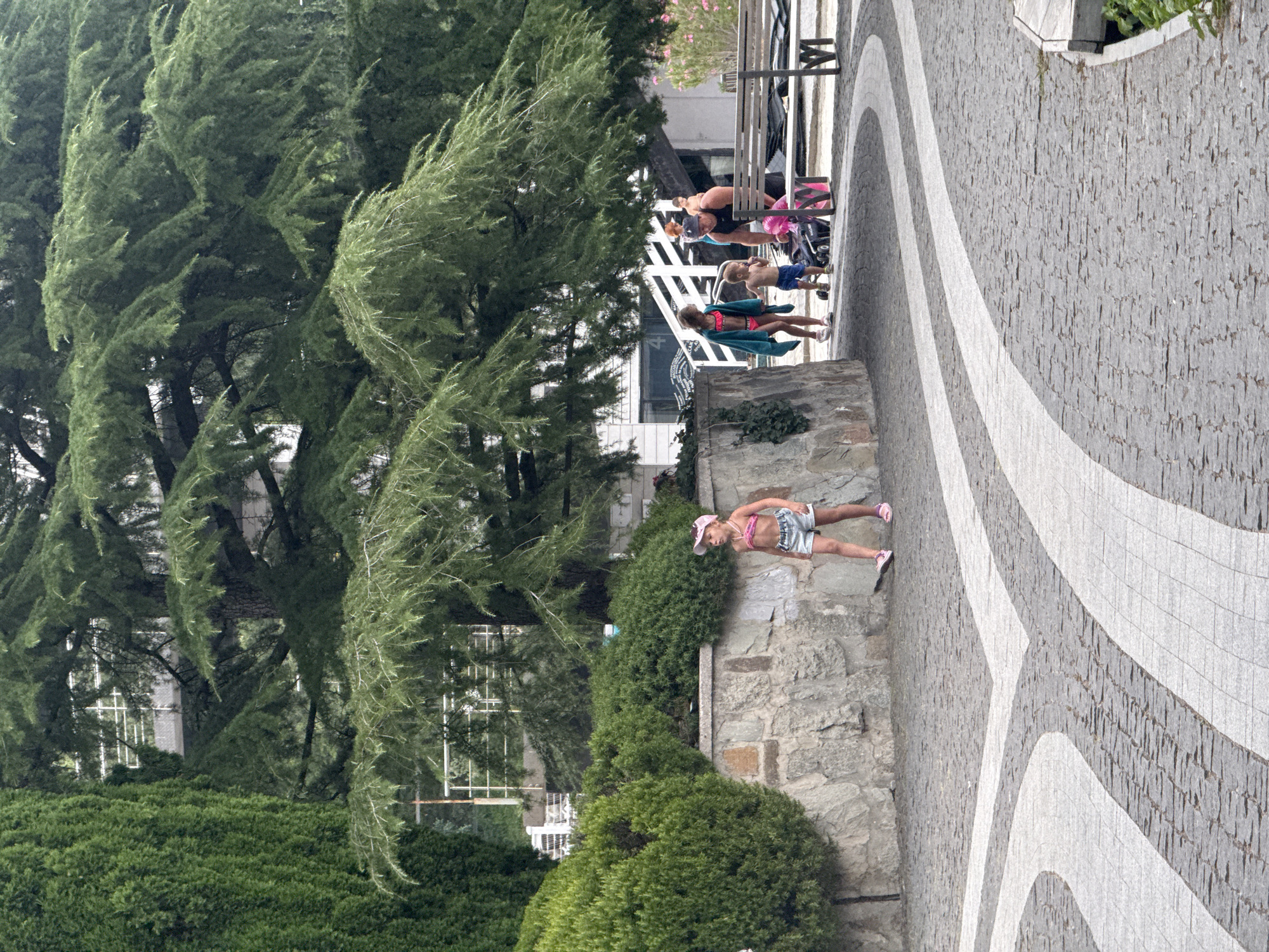 In this outdoor scene in Yalta, Ukraine, a young girl wearing a pink swimsuit and denim shorts stands on a stone wall. She has a pink hat on and is looking to her right. To her left, a woman in a dark top and pink shorts pushes a stroller. Further back, another child in a green swimsuit and a blue towel draped over their shoulders stands near a white structure. The background features large, green trees and a paved walkway with a distinctive pattern of gray and white lines. The time of day appears to be daytime, and the weather seems pleasant. The overall atmosphere suggests a leisurely outing. There is no visible text in the image.