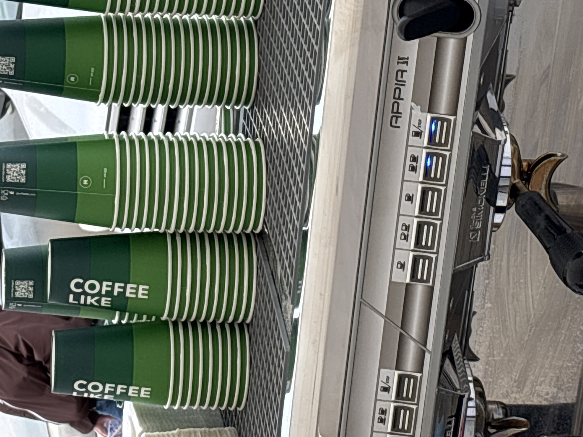 The image shows a stack of green and white "COFFEE LIKE" cups sitting next to an Appia II coffee machine. The cups are arranged neatly, and the coffee machine appears to be well-maintained. The image does not provide any visual indicators of the location within Perm, Russia. Without further information, a specific location within the city cannot be confirmed.