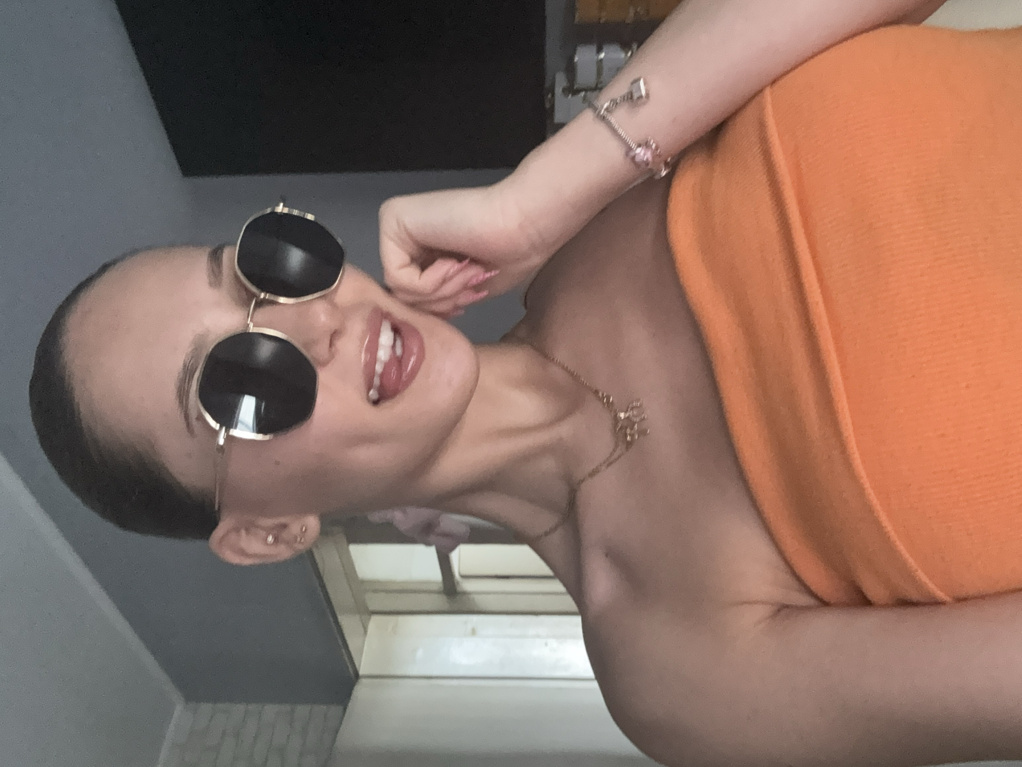 The image shows a young woman wearing sunglasses and an orange tube top. She has her hair pulled back and is sticking her tongue out playfully. She is also wearing necklaces and a bracelet. The background appears to be an indoor setting, possibly a home, with a wall and a doorway visible. There is no indication of the location being in the city of Sofia, Bulgaria.