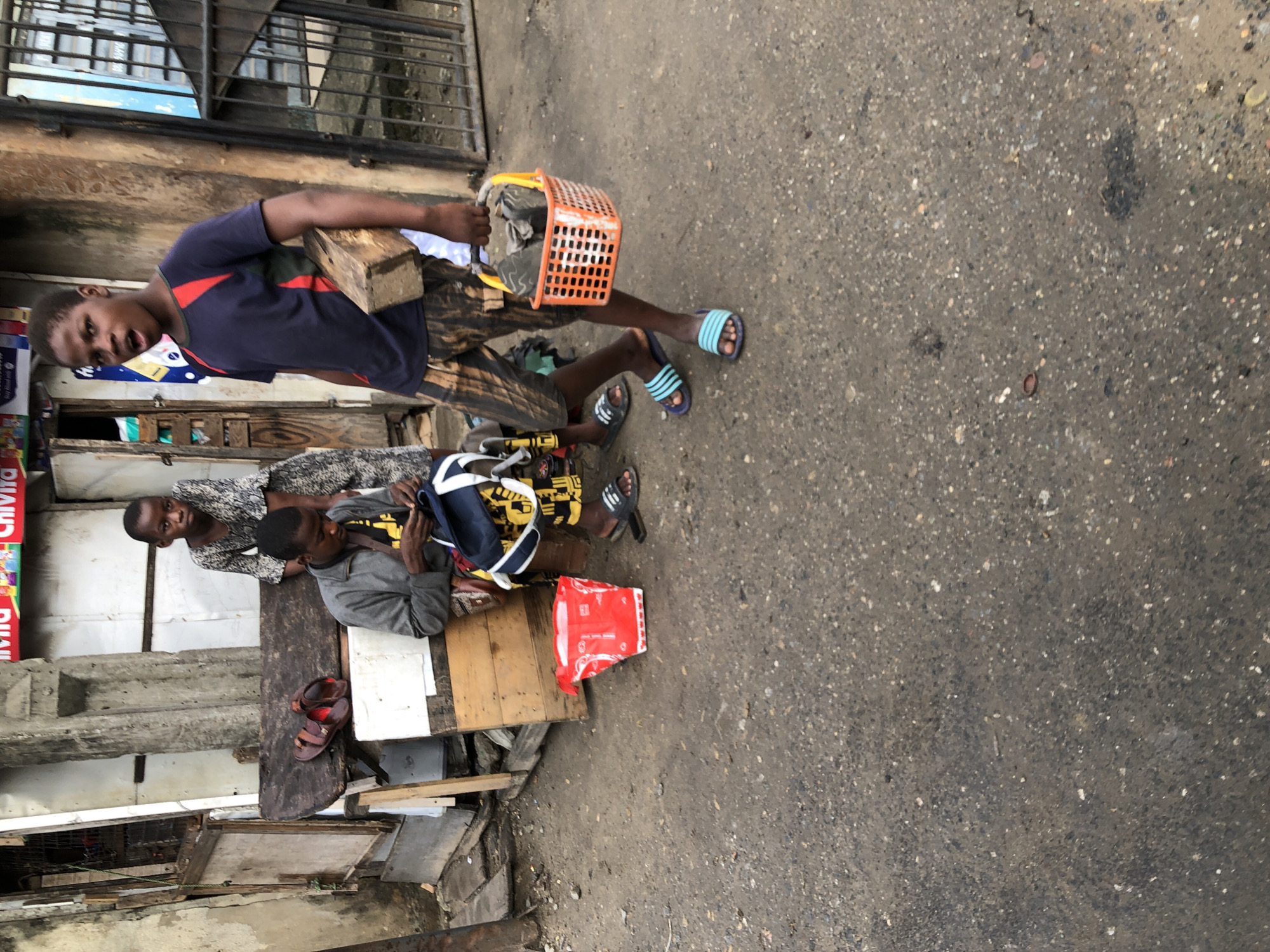 A group of children are in an urban outdoor setting, likely in Lagos, Nigeria. In the foreground, a young boy is walking towards the viewer, carrying a small orange basket with grey objects inside, possibly stones or building materials, and a wooden plank under his left arm. He is wearing a dark blue t-shirt with red accents and patterned shorts. Behind him, to the left, another child is seated on a wooden bench, sewing or mending something with a blue bag beside them. They are wearing a grey jacket and patterned trousers. To the far left, a younger child stands near the seated child, wearing a dark patterned dress. In front of the seated child is a bright red bucket. The environment appears to be a street with worn asphalt ground, partially constructed buildings with debris scattered around, and a metal gate or fence in the background. The scene depicts everyday life in a busy urban area.
